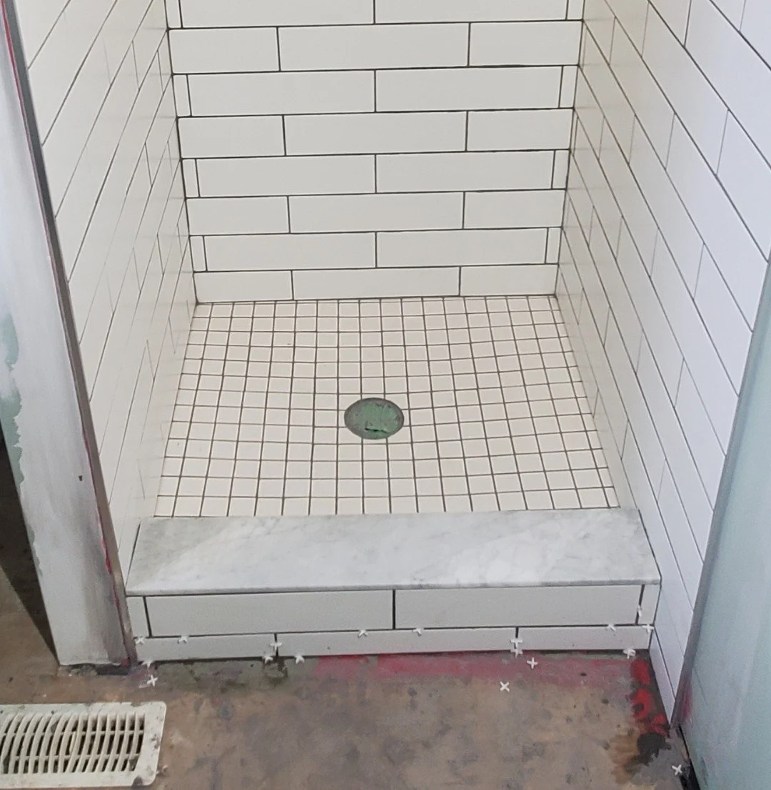 Tile shower with white subway tiles on the walls, small square tiles on the floor, a shiny round drain, and a marble threshold at the entrance.