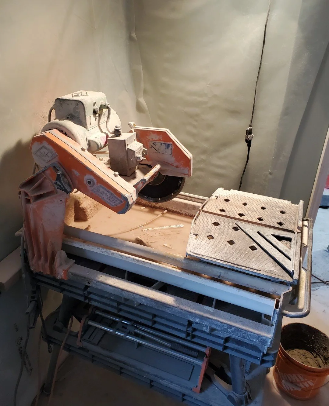 A dusty miter saw on a workbench in a construction or renovation site with a bucket of concrete nearby.