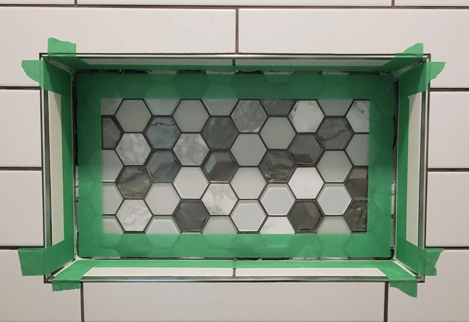 A tile work in progress with hexagonal tiles in shades of white, gray, and black, surrounded by green painter's tape on a tiled wall.