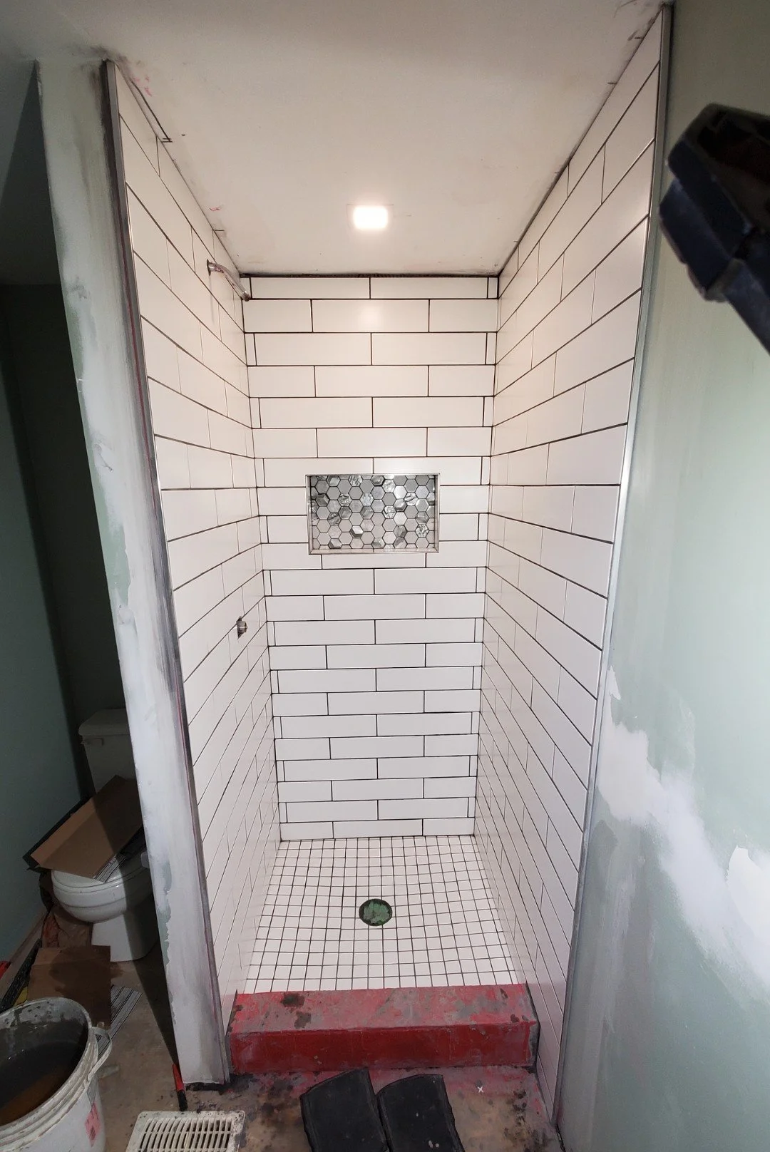 Inside a shower stall with white subway tile walls, a small hexagon tile niche, and a tiled floor with a central drain. The stall is under construction with visible unfinished edges and construction materials nearby.