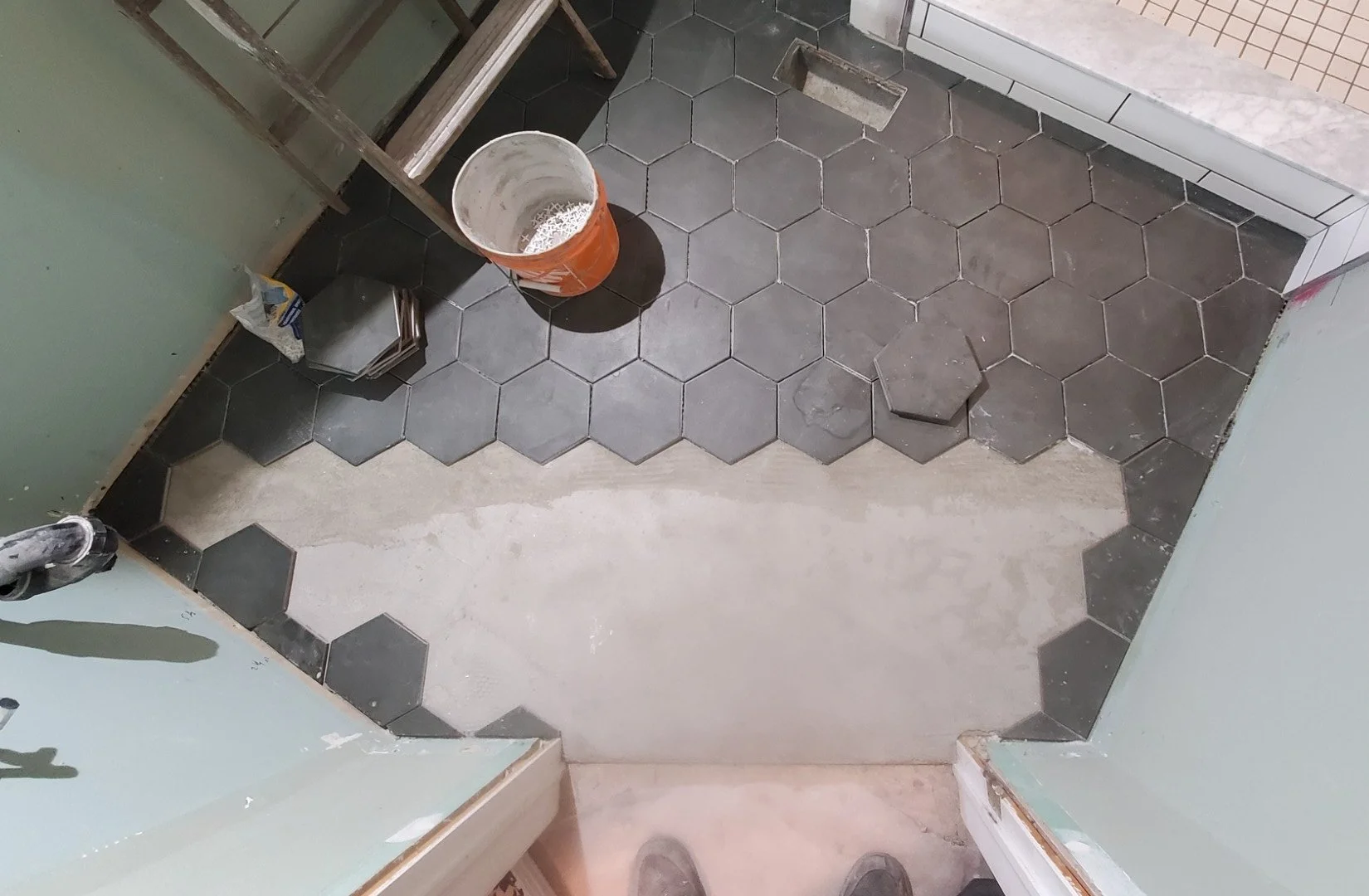 Room under construction with black hexagonal floor tiles partially installed, a bucket, tools, and shoes visible.