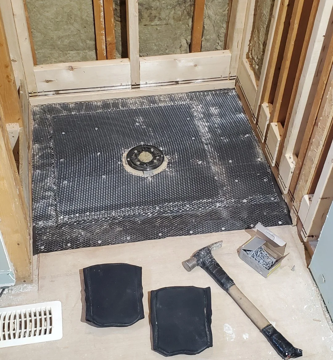 A bathroom under construction with a shower area covered in a black waterproof membrane, wooden framing, and a drain in the center. There are two pads, a hammer, and a box of nails or screws on the floor.