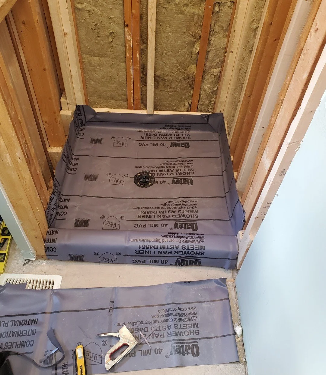 A shower pan installed in a bathroom under construction, with the drain in place, surrounded by unfinished walls and construction tools on the floor.