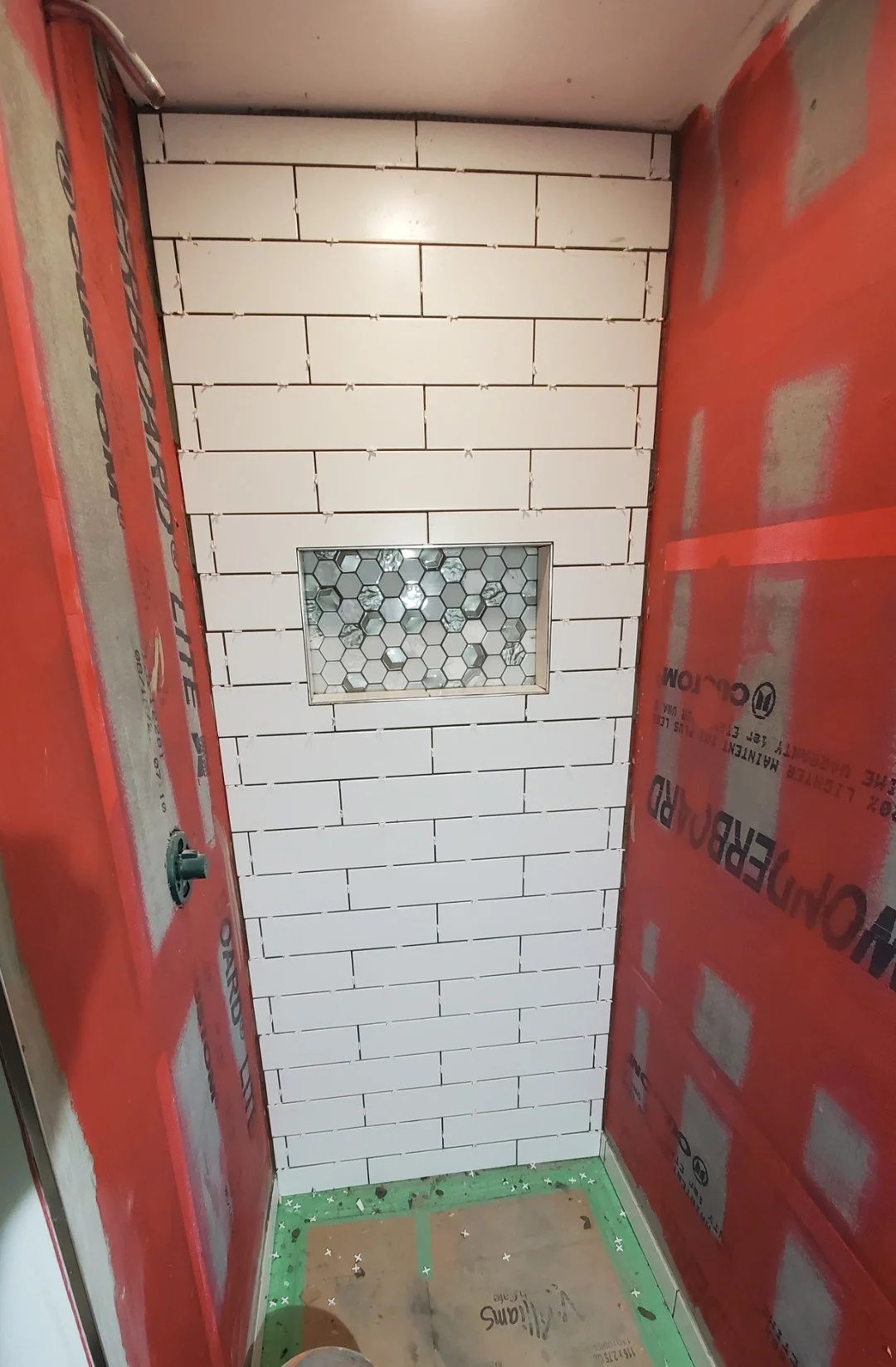 Construction of a small shower with white subway tiles on the wall, a small hexagonal tile niche, unfinished walls, red waterproof barrier on the sides, and green protective covering on the floor.