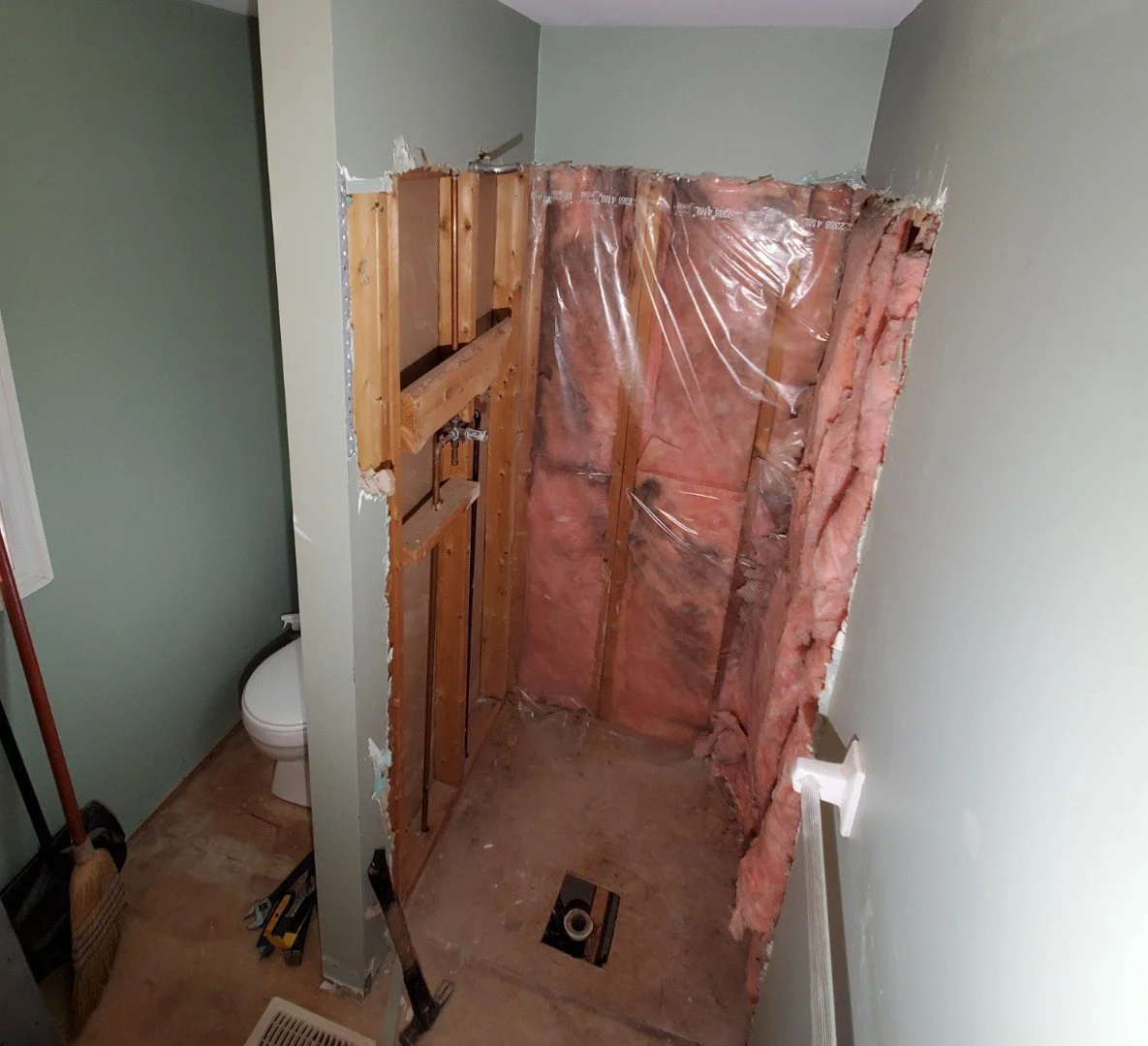 A bathroom undergoing renovation with a partially demolished wall, exposing wooden studs, pink insulation, and plastic sheeting, with tools on the floor.