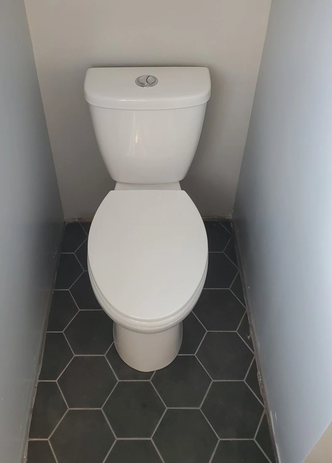 A white toilet in a small bathroom with gray walls and black hexagonal floor tiles.