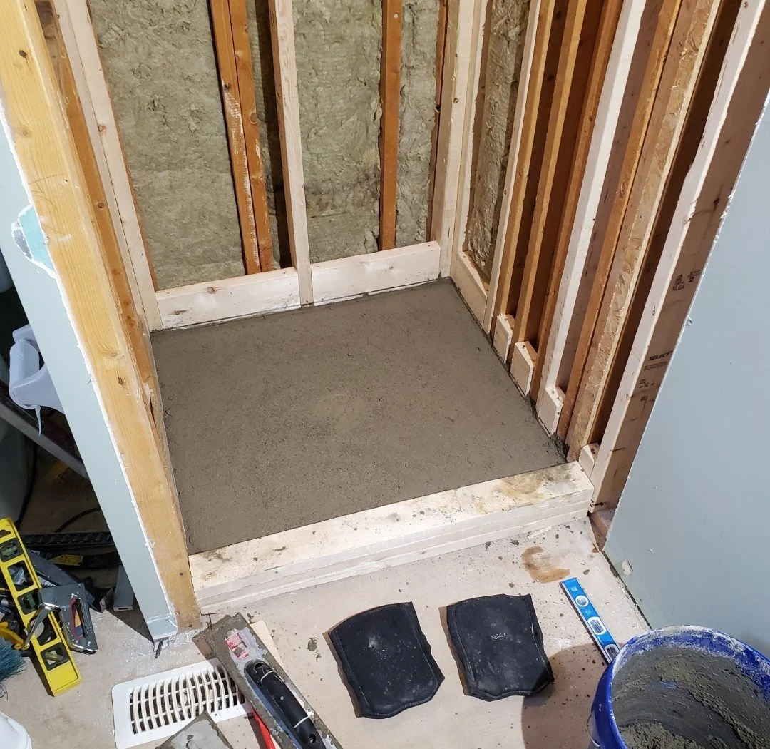 A bathroom under construction with a newly poured concrete slab floor, unfinished wall framing, and construction tools including a spirit level, black rubber mats, and a bucket of concrete.