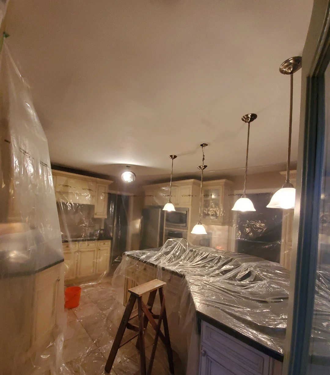 Kitchen under renovation covered with plastic sheets and a stool, with pendant lights hanging from the ceiling.