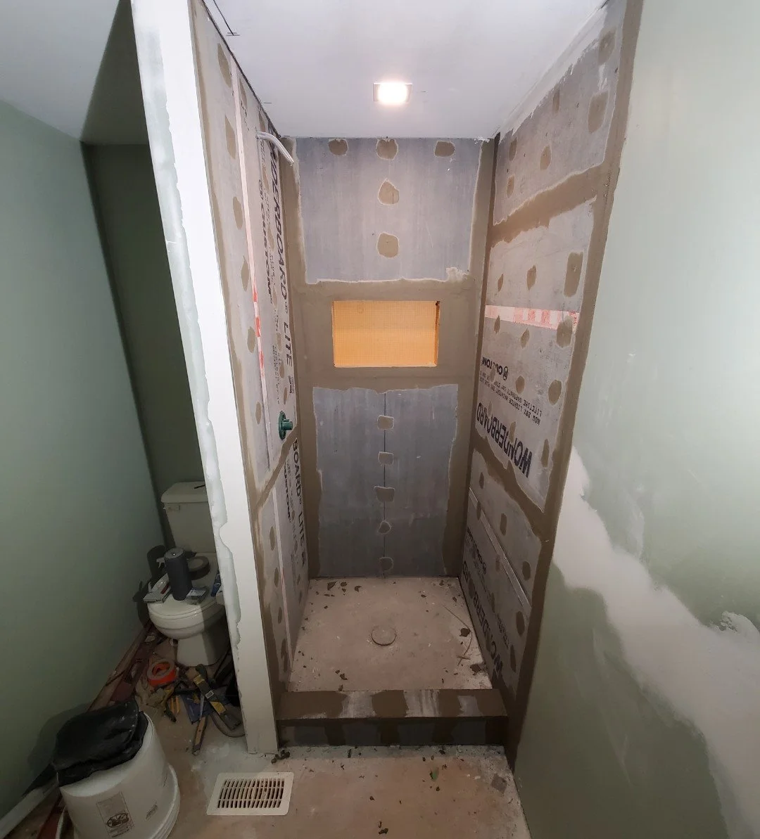 A small bathroom under renovation with an unfinished shower area, partly painted green walls, and construction tools and materials on the floor.