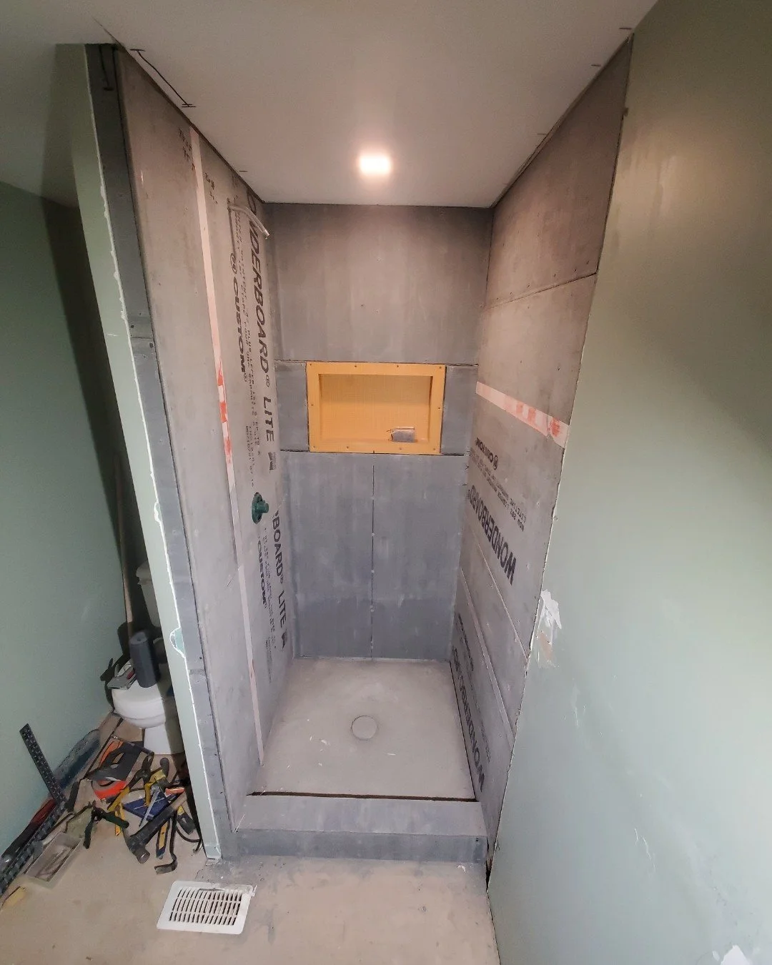 Bathroom under construction with a shower stall installed, unfinished walls, tools on the floor, and a small yellow access panel on the back wall.
