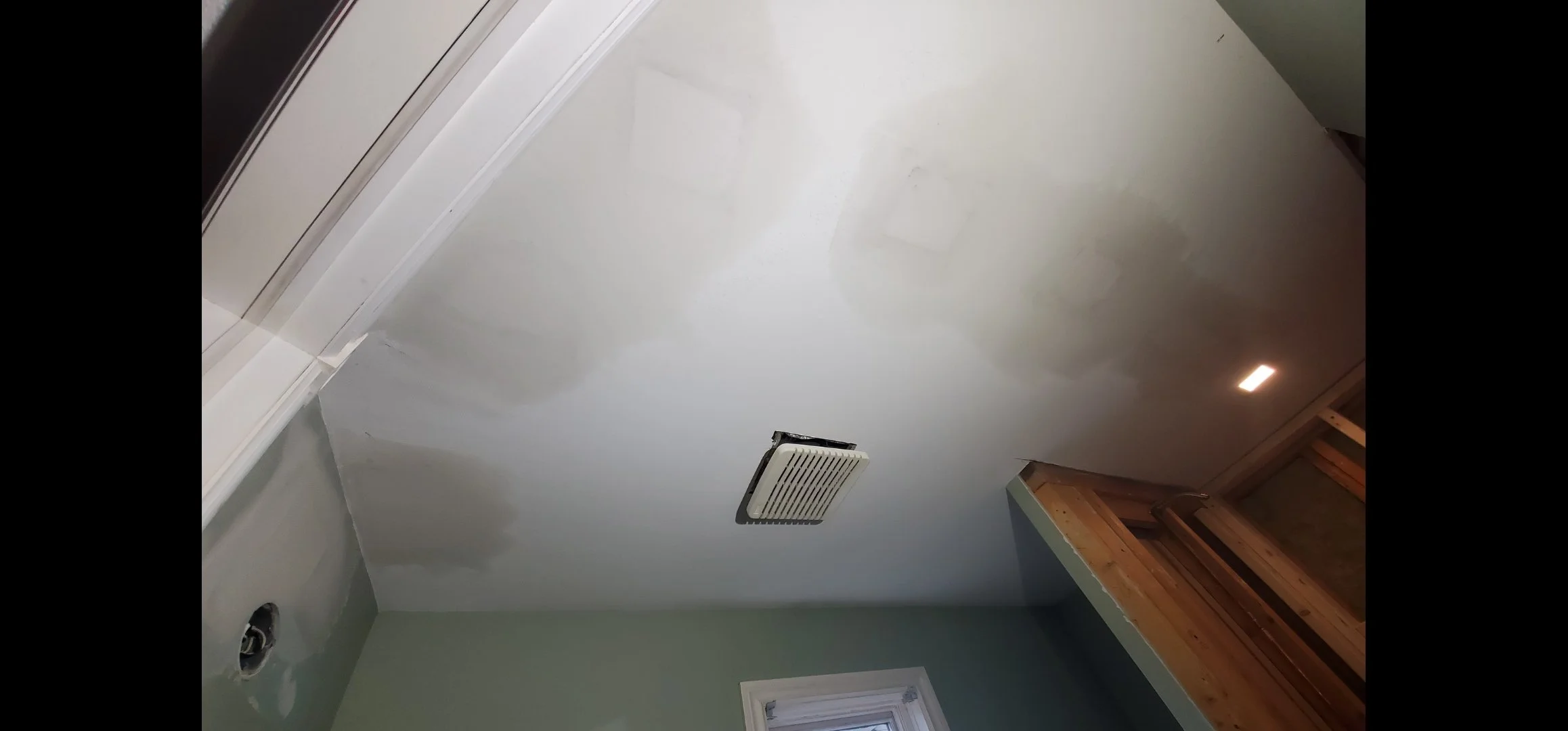 Ceiling of a room under construction with a vent and a light fixture, and an unfinished corner with exposed wood framing.