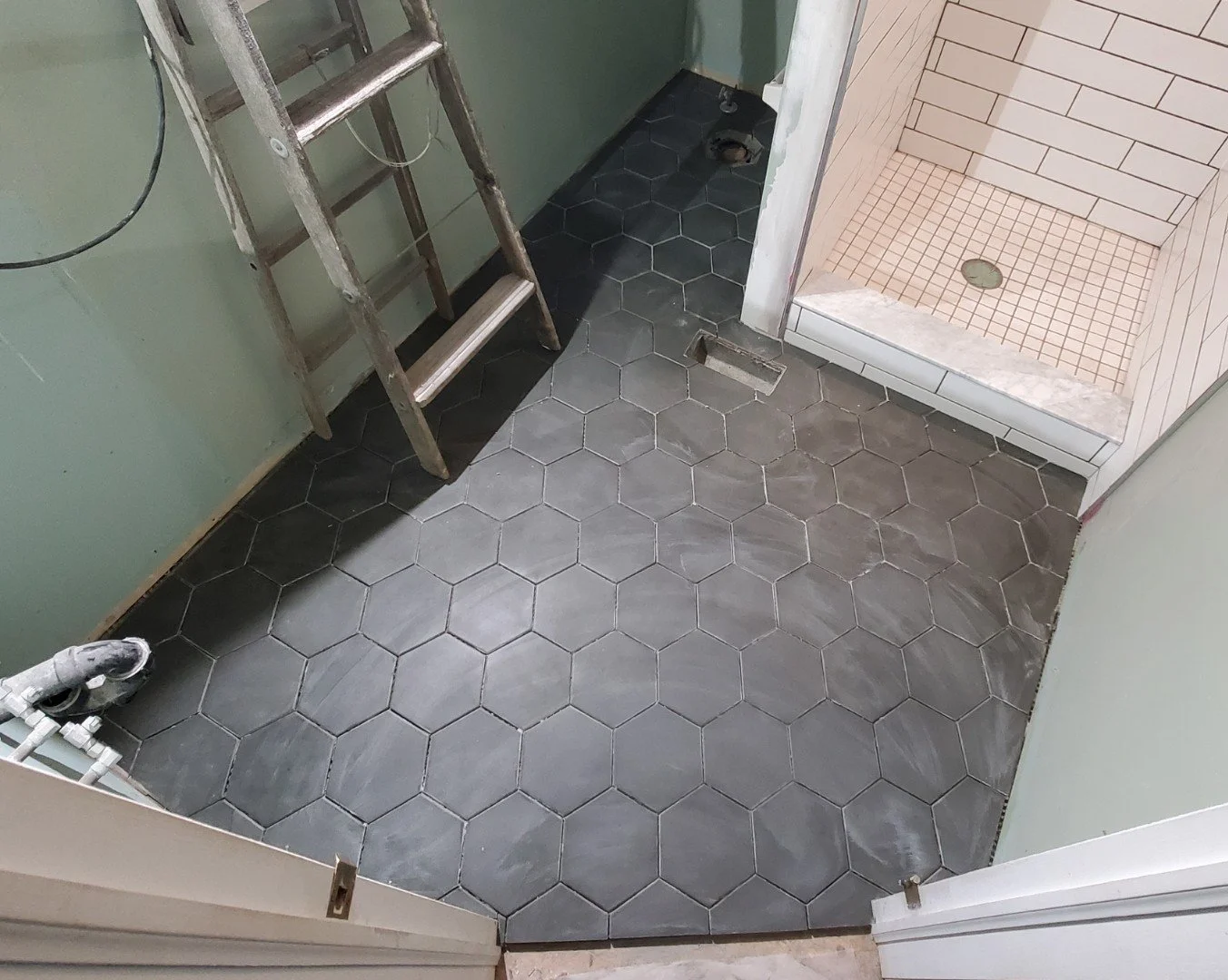 Bathroom under renovation showing dark hexagonal floor tiles, a ladder, and a small shower stall with light-colored tiles.