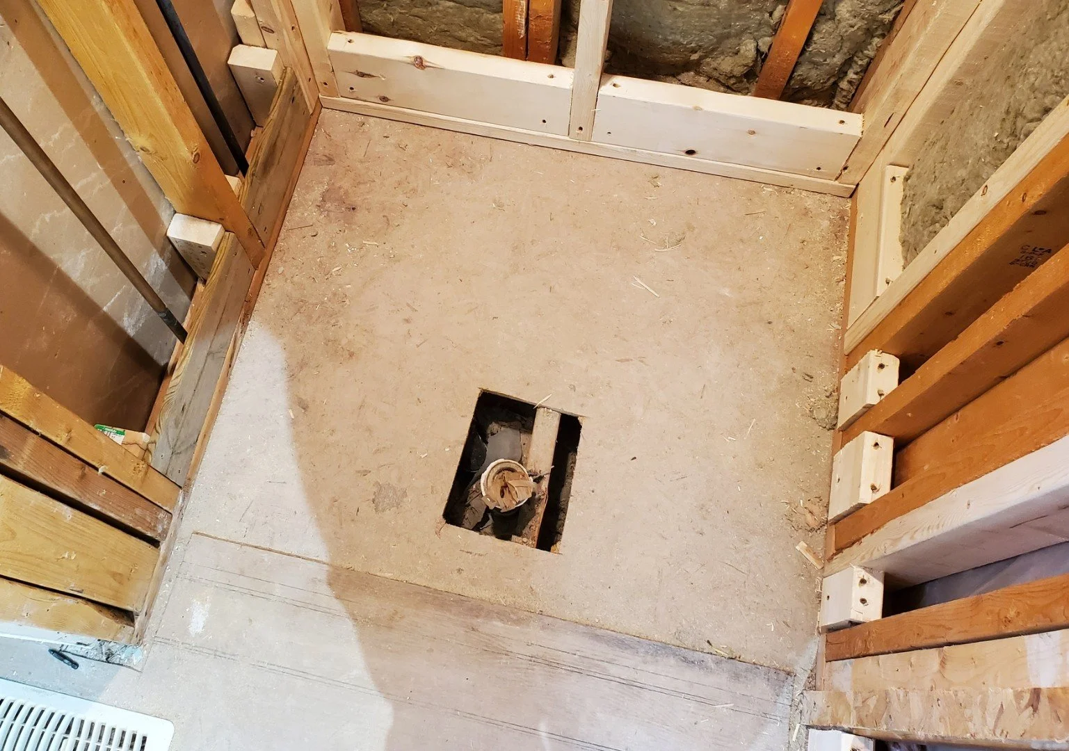 Interior of a room under construction, with exposed framing and a hole in the floor for plumbing or electrical work.
