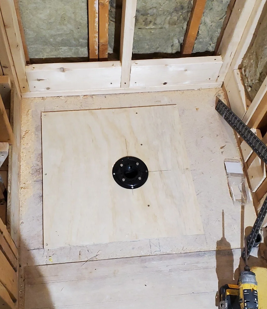 Construction site with a wooden floor and a black circular fixture in the center, surrounded by unfinished wooden framing and wall insulation.