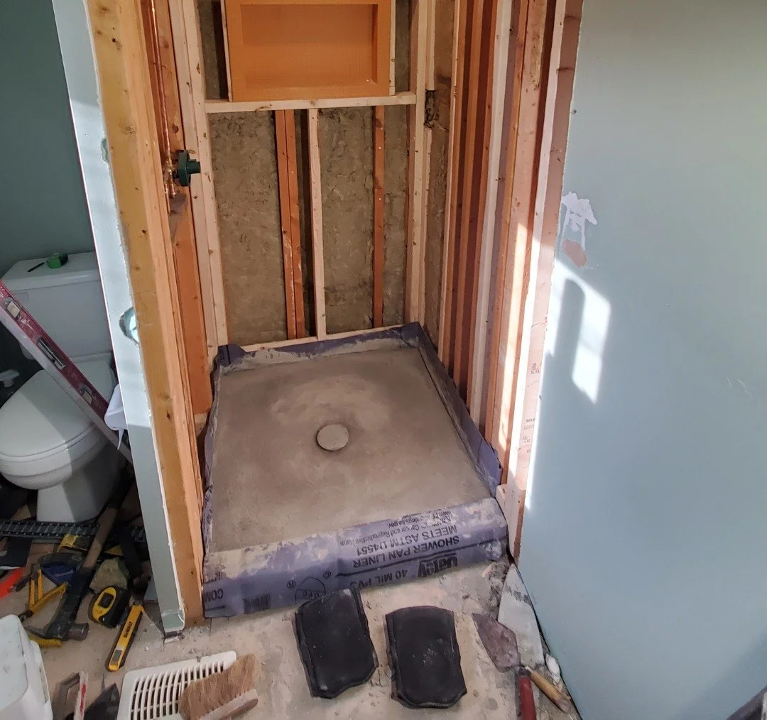 Empty shower base with plumbing, isolated walls, and construction tools surrounding it in a bathroom under renovation.