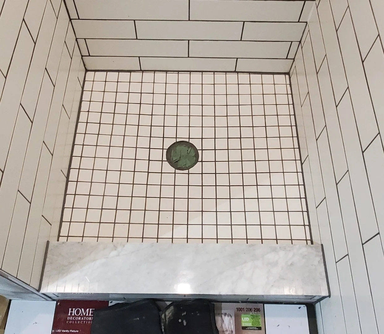 Empty shower stall with small square beige tiles on the floor and rectangular tiles on the walls, with a round drain in the center.