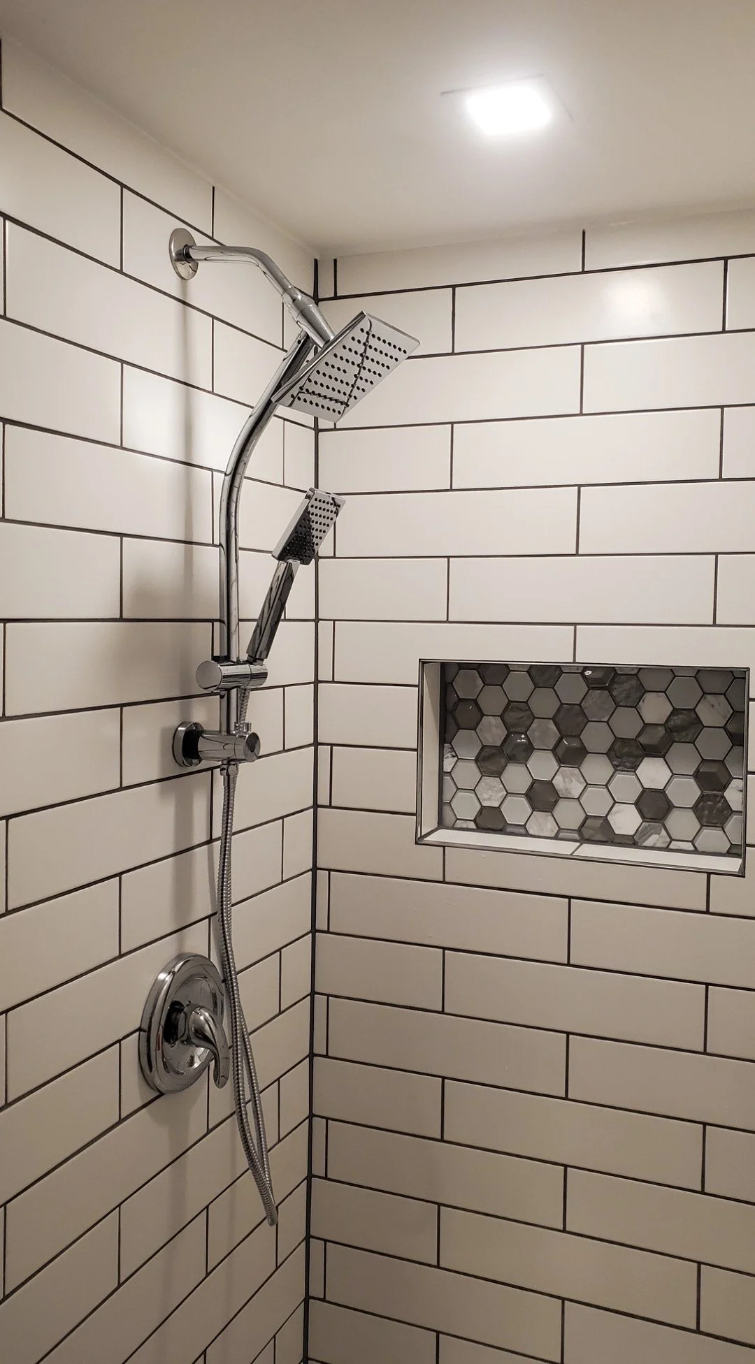Shower with white subway tiles on the wall, two black-dotted showerheads, a built-in niche with hexagon tiles, and a ceiling light.