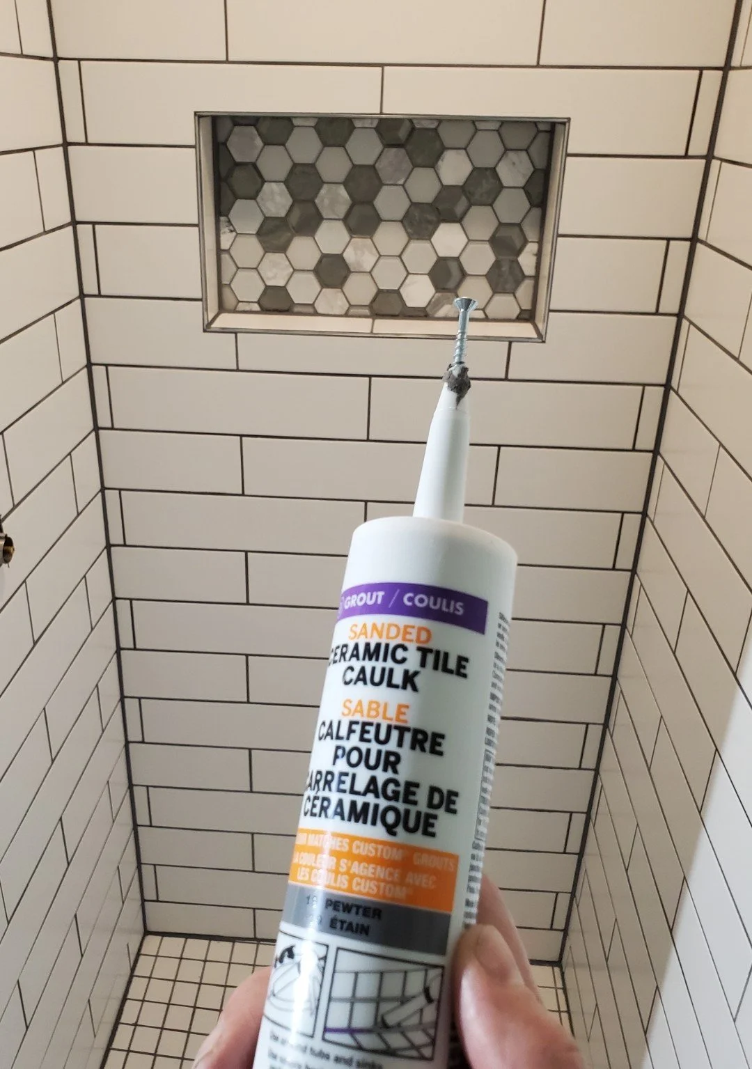 A hand holding a tube of sanded ceramic tile caulk in front of a tiled bathroom shower with a recessed niche.