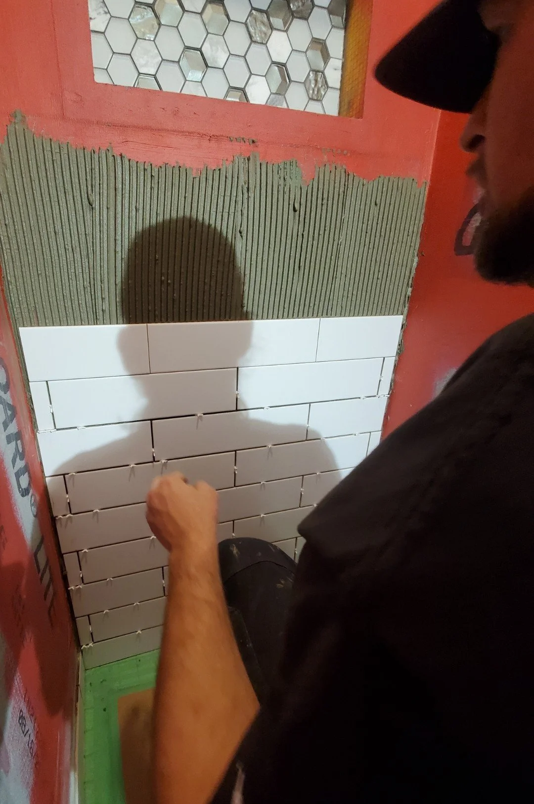 A man is tiling a wall with white rectangular tiles in a partially finished room. The wall has an upper section with greenish tiles and a small mirror or window above. The man is wearing a black cap and black shirt, working on the lower part of the w