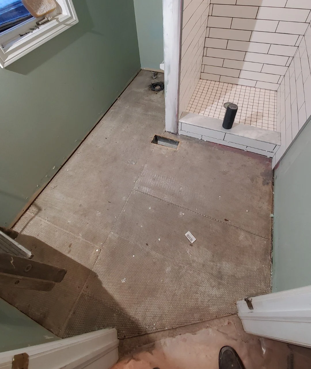 A bathroom under renovation showing a partially removed floor, with a shower area on the right that has tiled walls and a black pipe on the floor, and a green wall with a window on the left.