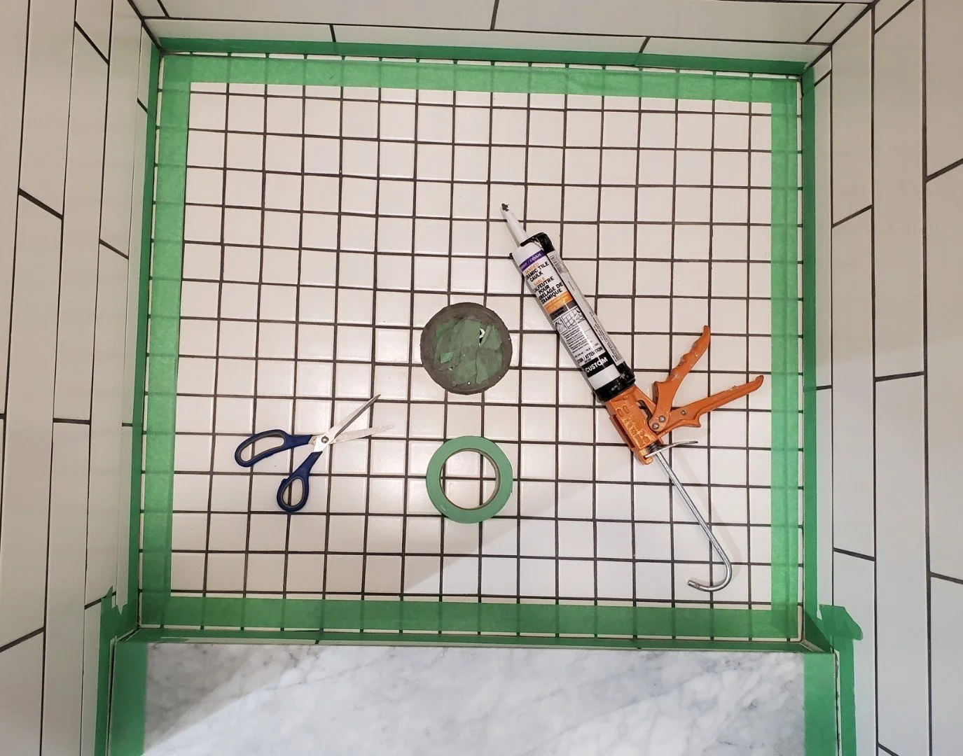 Tools for tiling a shower stall, including scissors, green painter's tape, a tube of silicone sealant, and a caulking gun, are arranged inside a tiled shower stall with a partially completed green border.