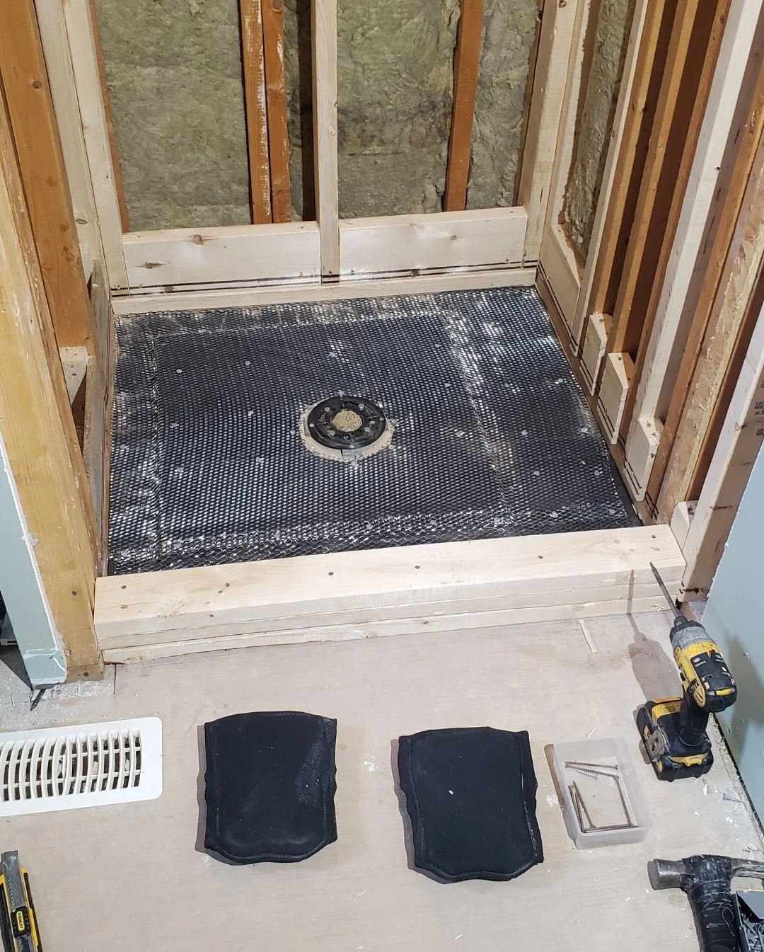 Half-completed shower stall under construction with a black waterproof membrane, a drain in the center, and wooden framing. Tools and materials are visible nearby.