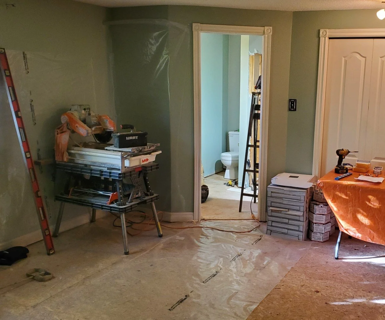 Interior of a room under renovation with construction tools, a ladder, a work table, and a visible bathroom through an open doorway.