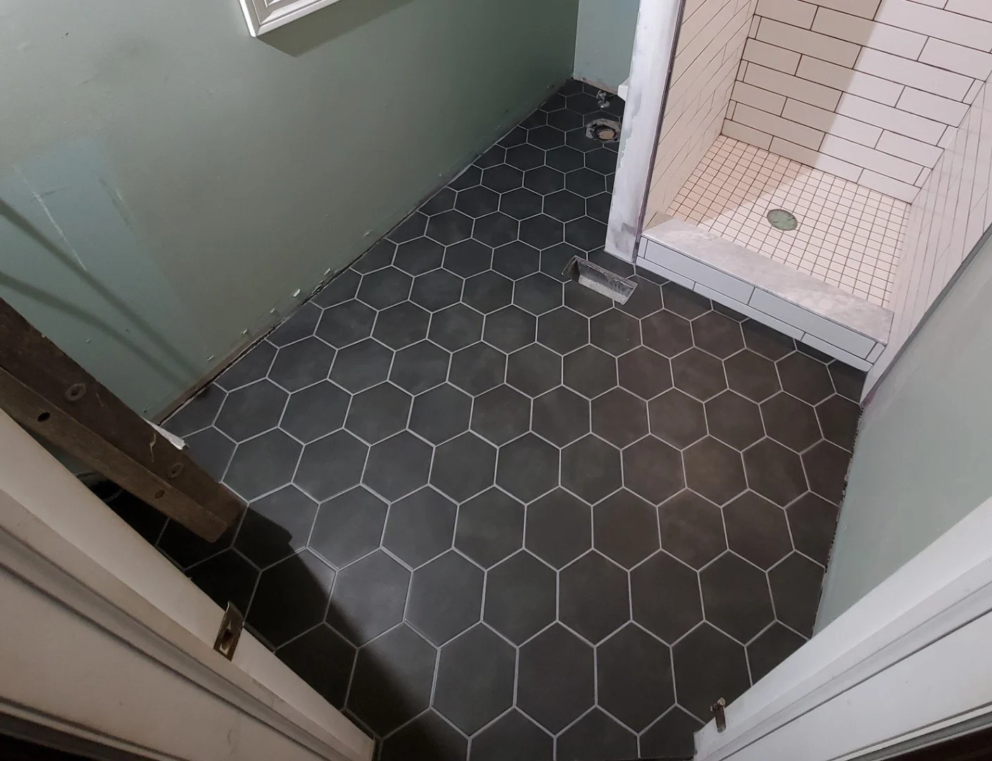 Bathroom floor with black hexagon tiles, shower with white square tiles, and green painted walls. Shower drain visible, and part of doorway with partial wall framing.