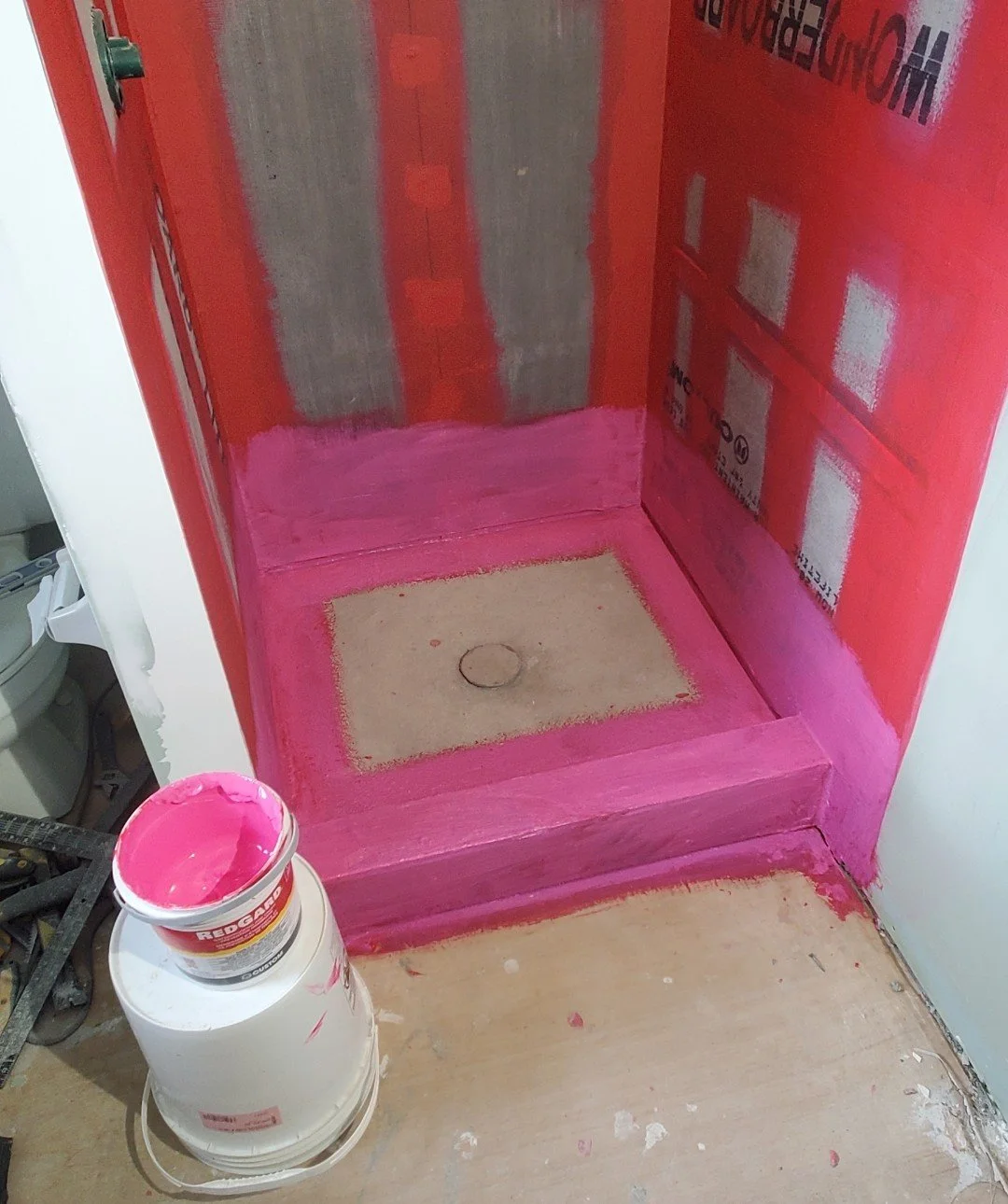 A shower base or small alcove with partially painted walls in pink primer. A can of pink paint and painting supplies are on the floor nearby. The walls are covered with red fireproof drywall with labels, and the bottom of the walls has a layer of pin