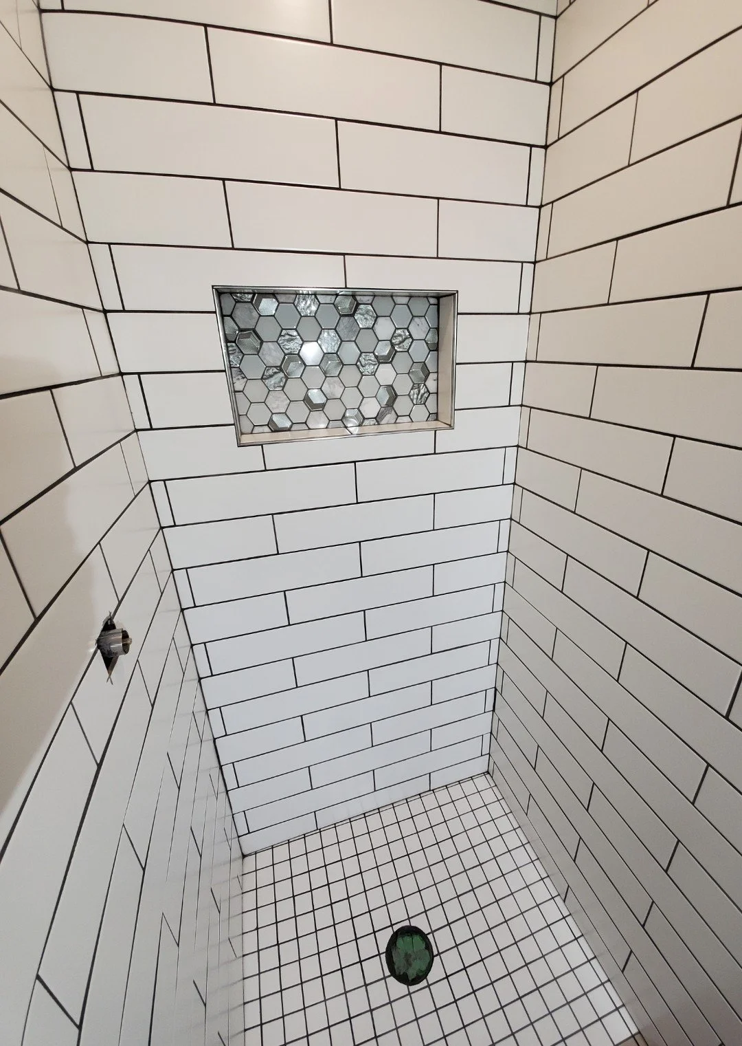 A tiled shower with white subway tiles on the walls, smaller square tiles on the floor, a small hexagon tile window, and a round drain on the floor.