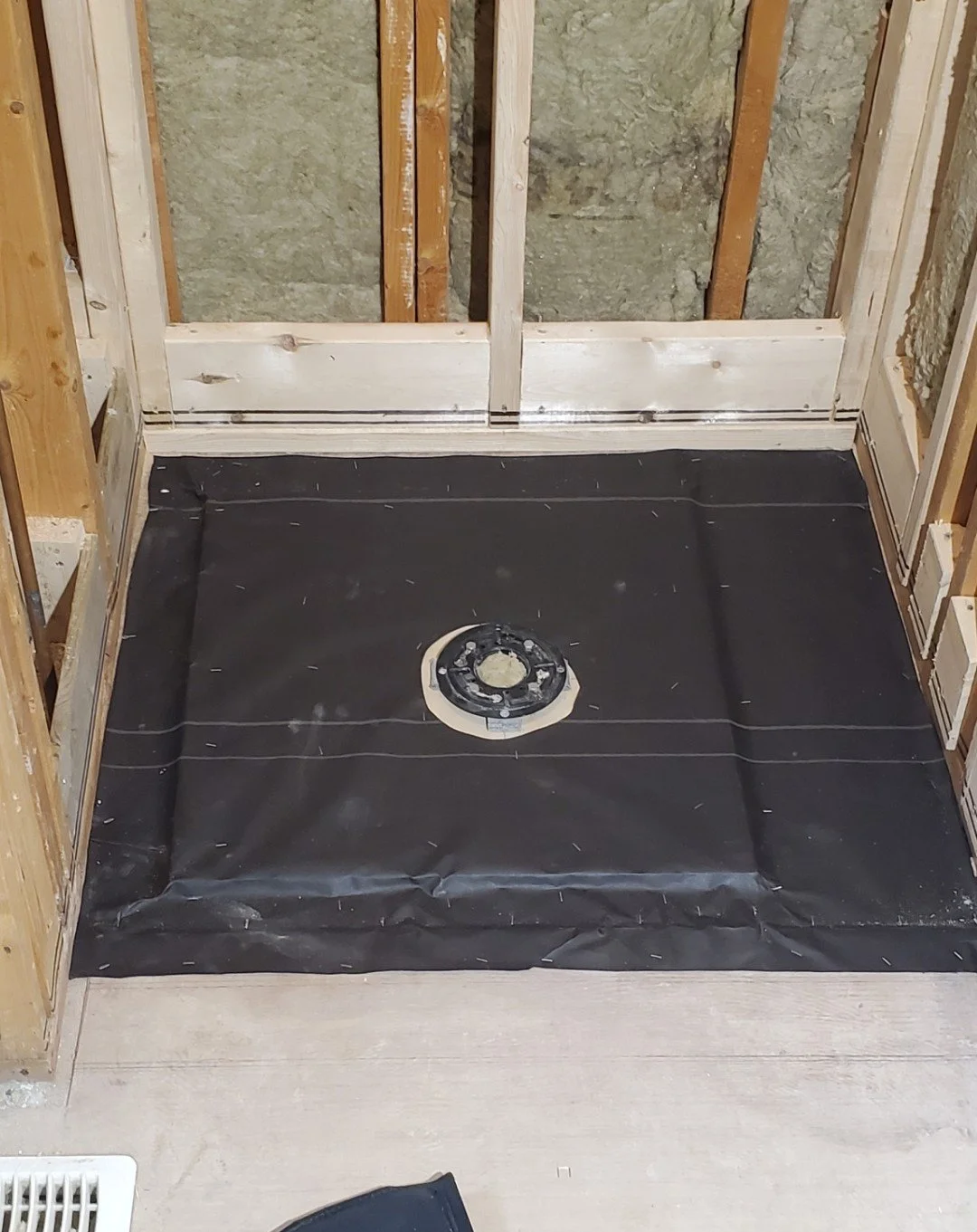 Bathroom floor with black waterproofing membrane and a drain installed, surrounded by unfinished wooden wall studs.
