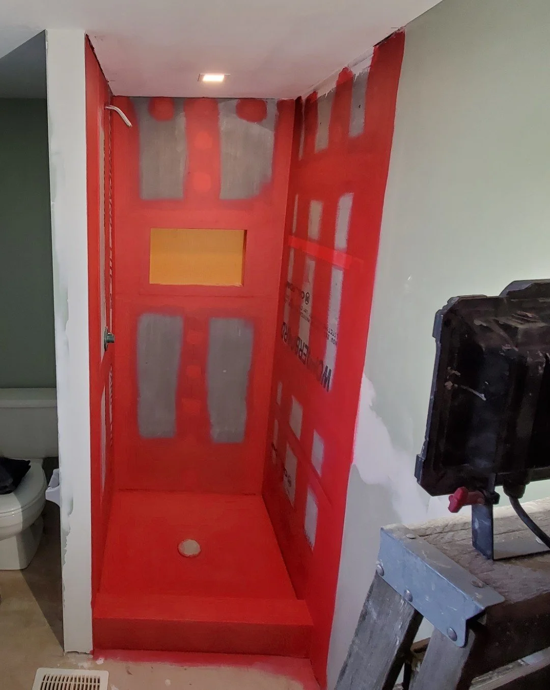 This image shows the interior of a shower stall under construction with red waterproofing membrane applied to the walls and floor. There is a showerhead on the left wall and a drain hole in the center of the shower floor.
