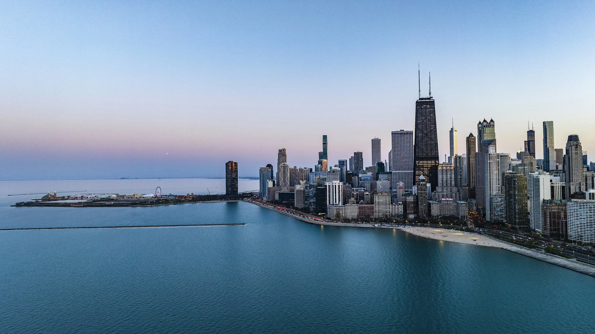 Aerial view of Chicago skyline at dusk with Lake Michigan in the foreground, including iconic skyscrapers like John Hancock Center and Navy Pier.