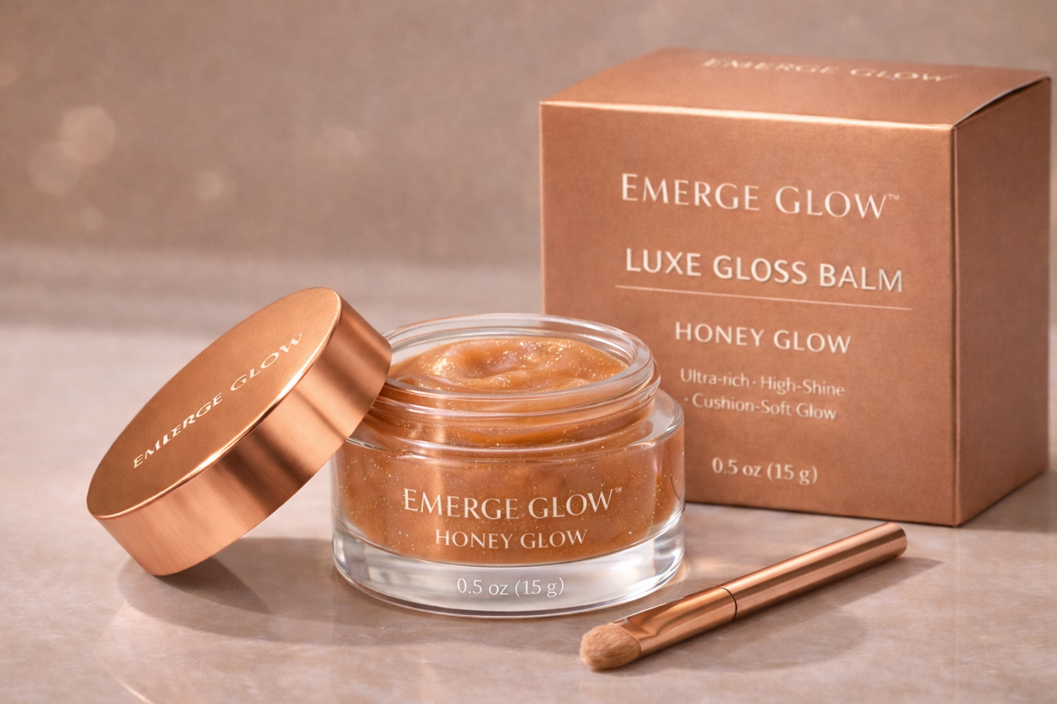 Open jar of Emerge Glow Honey Glow Luxe Gloss Balm with gold lid, alongside its packaging box, a small makeup brush, all on a beige surface.