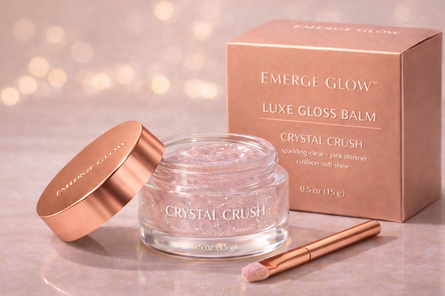 A cosmetic glitter balm in a clear jar with pink and iridescent glitter, a rose gold lid labeled "EMERGE GLOW," a pink box labeled "Emerge Glow Luxe Gloss Balm Crystal Crush," and a rose gold applicator brush, on a soft pink surface with blurred ligh