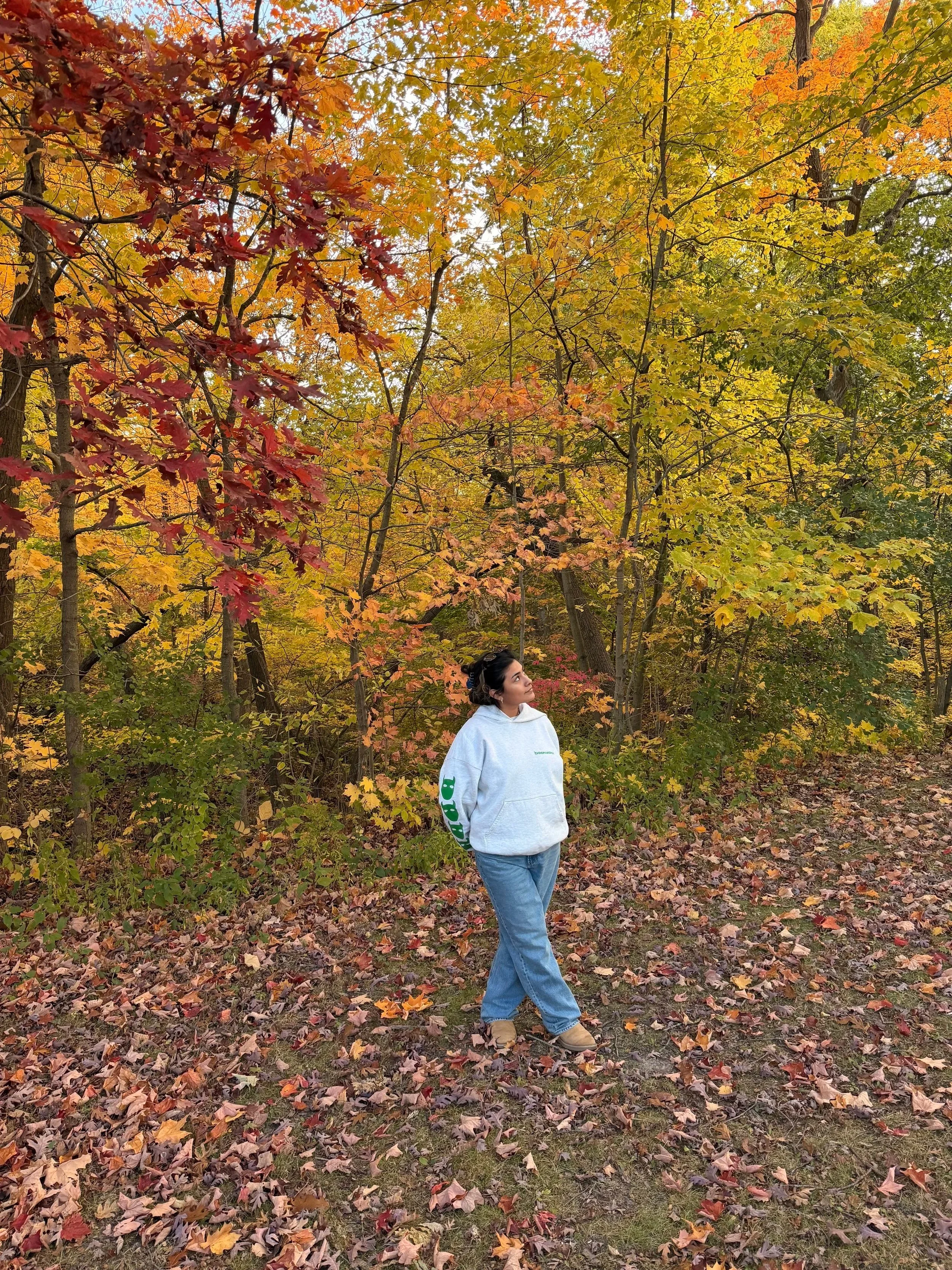 A woman in a white hoodie and blue jeans standing on a forest trail covered with fallen leaves, surrounded by trees with autumn foliage in shades of red, orange, and yellow.