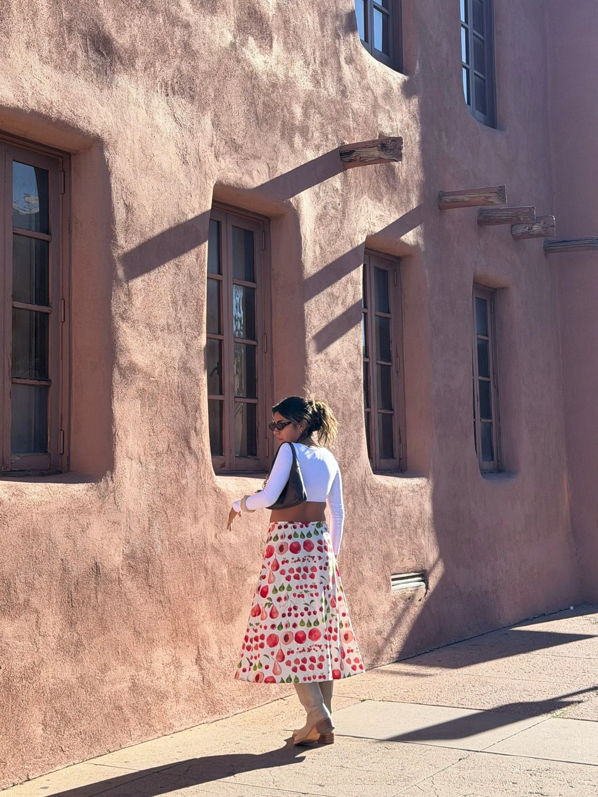 A woman standing in front of a pink stucco building with multiple windows and wood beams protruding from the wall casting shadows, wearing sunglasses, a white long-sleeve top, a colorful skirt with fruit patterns, and carrying a black handbag.