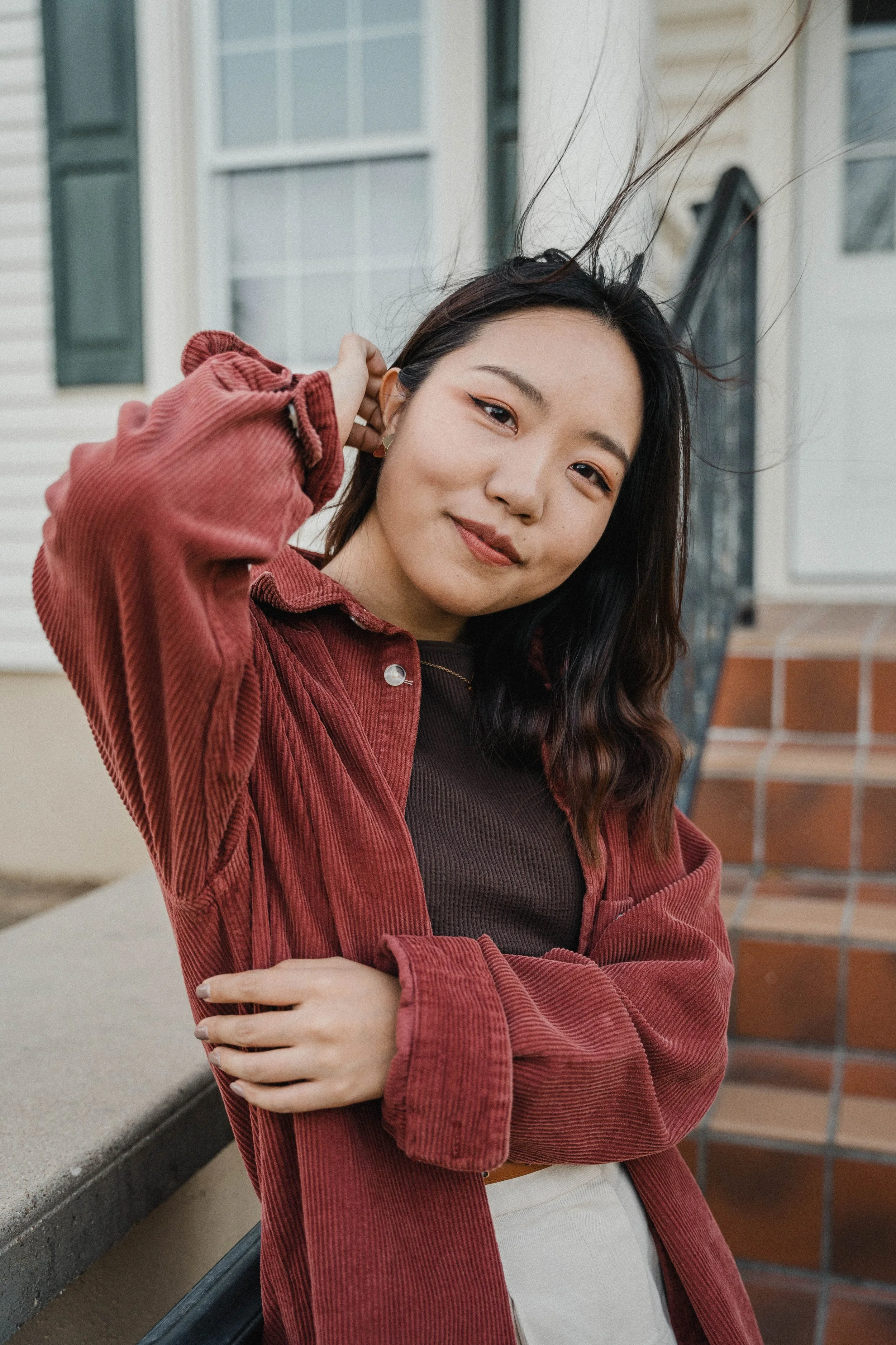 A woman with long brown hair, wearing a striped beige and white jacket, is sitting sideways on a wooden chair with metal frame, in front of a beige backdrop with studio lighting equipment visible.