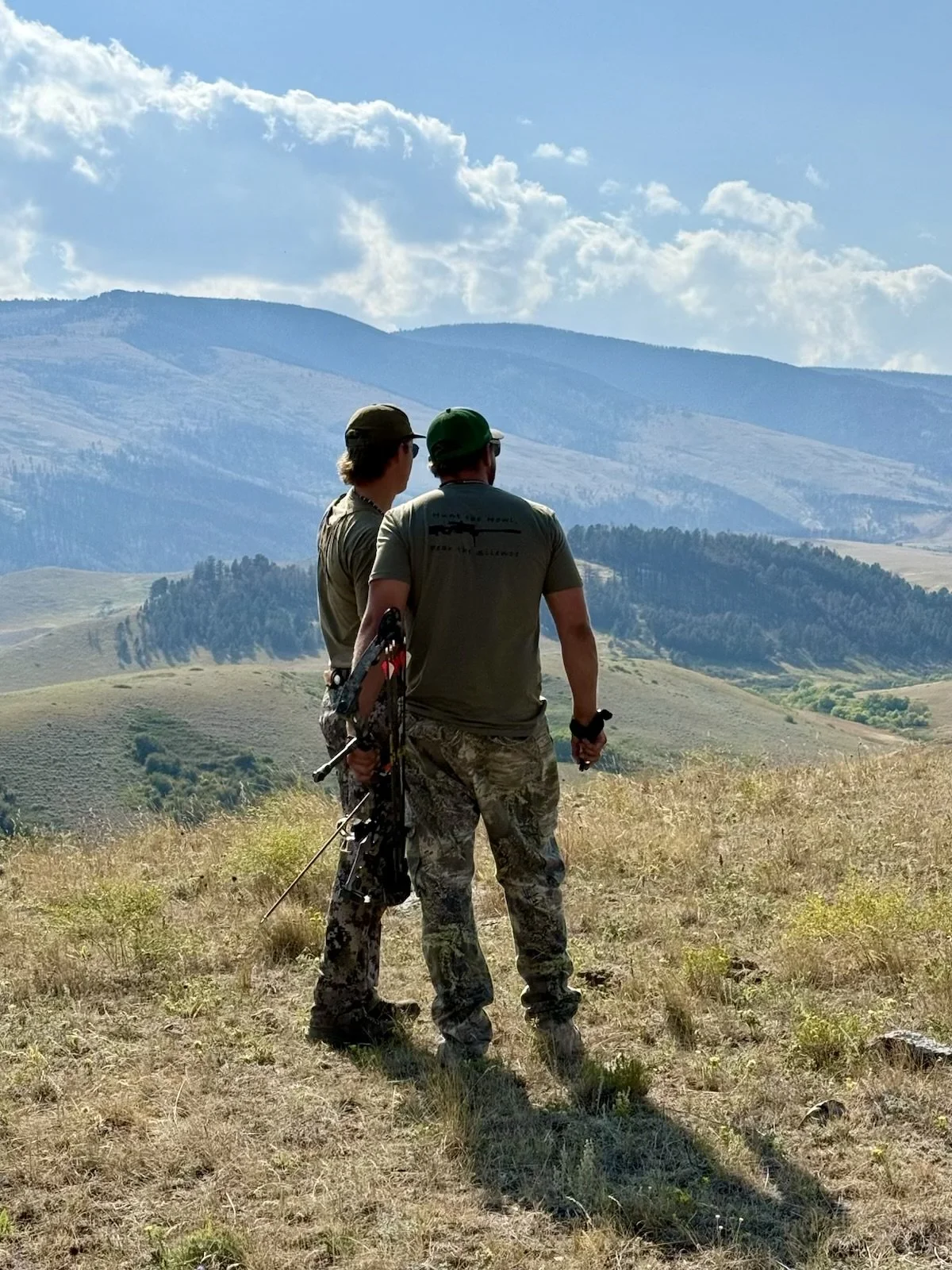 Two people standing in a grassy field with mountains in the background, one holding a bow and quiver of arrows, dressed in camouflage pants and T-shirts, both wearing caps, under a partly cloudy sky.