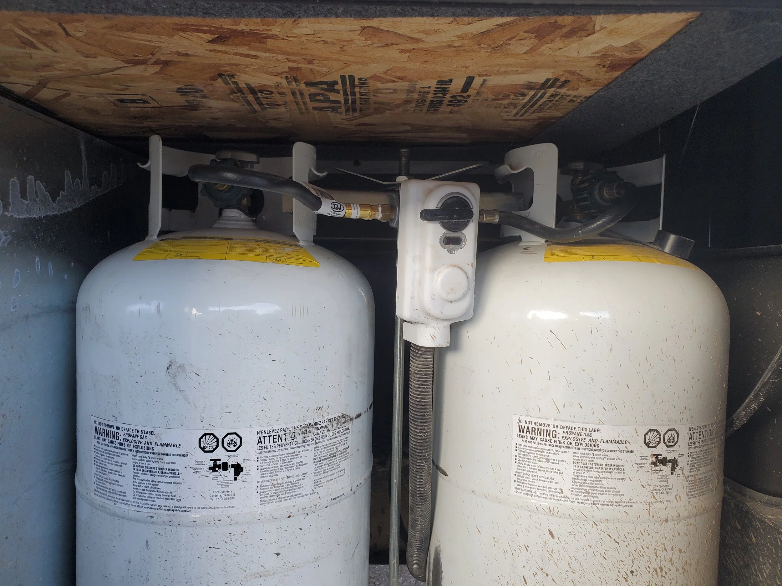 Two propane tanks connected to a pipeline under a wooden shelf
