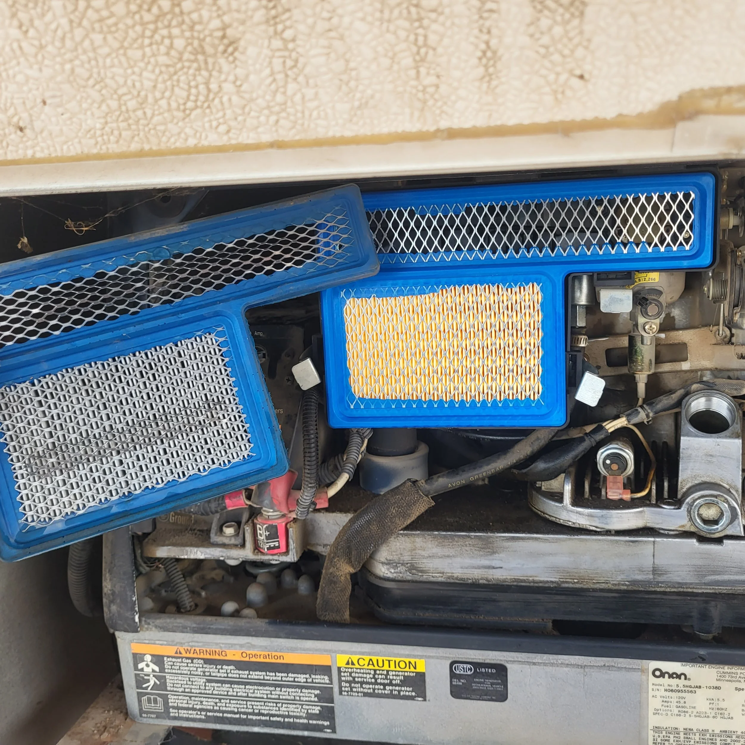 Close-up of an engine with three blue and yellow air filters mounted on top, with various mechanical components and wiring visible underneath.