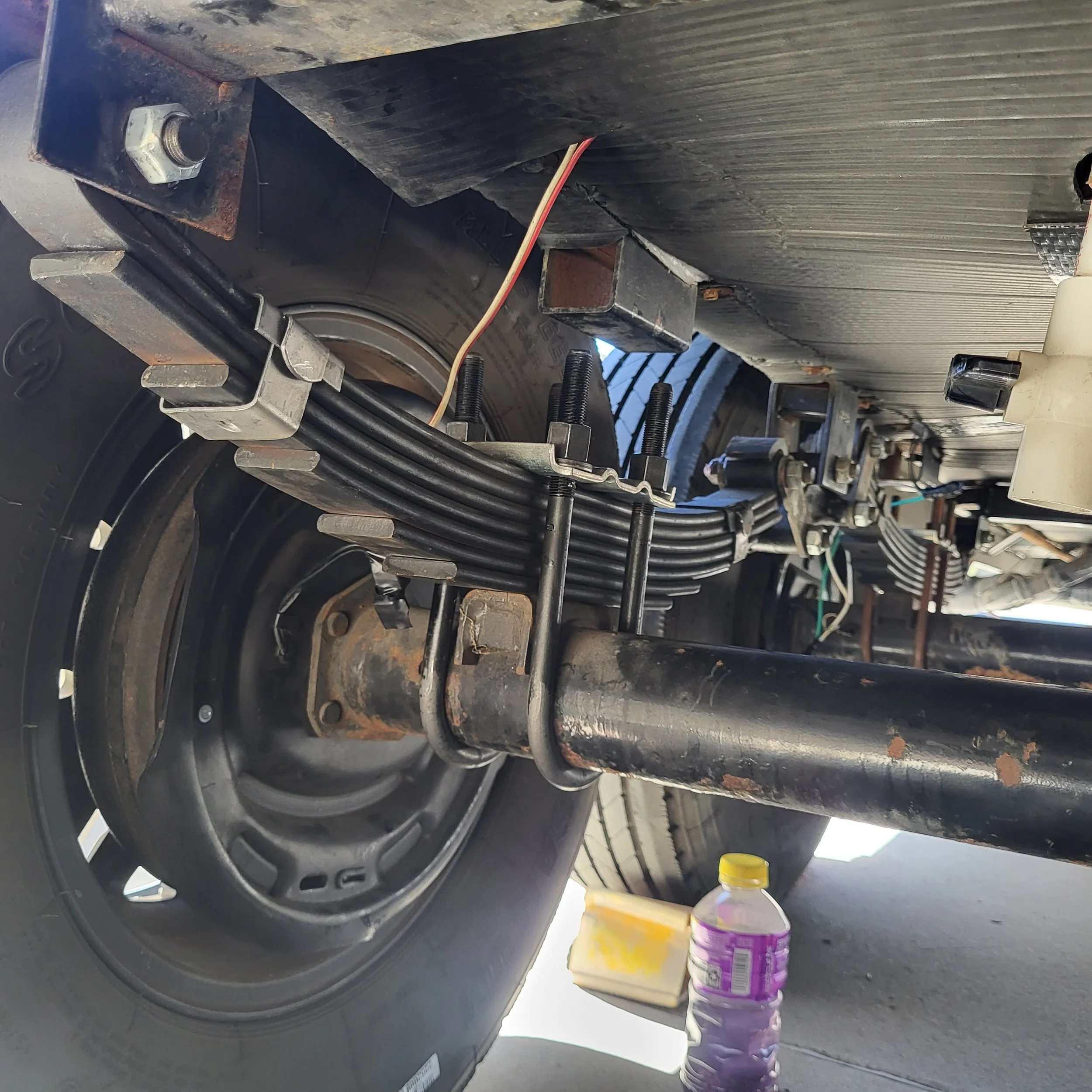Close-up of a large vehicle's wheel and suspension system showing multiple black cables, metal brackets, and bolts, with a purple water bottle, a container, and a yellow scrub pad underneath.