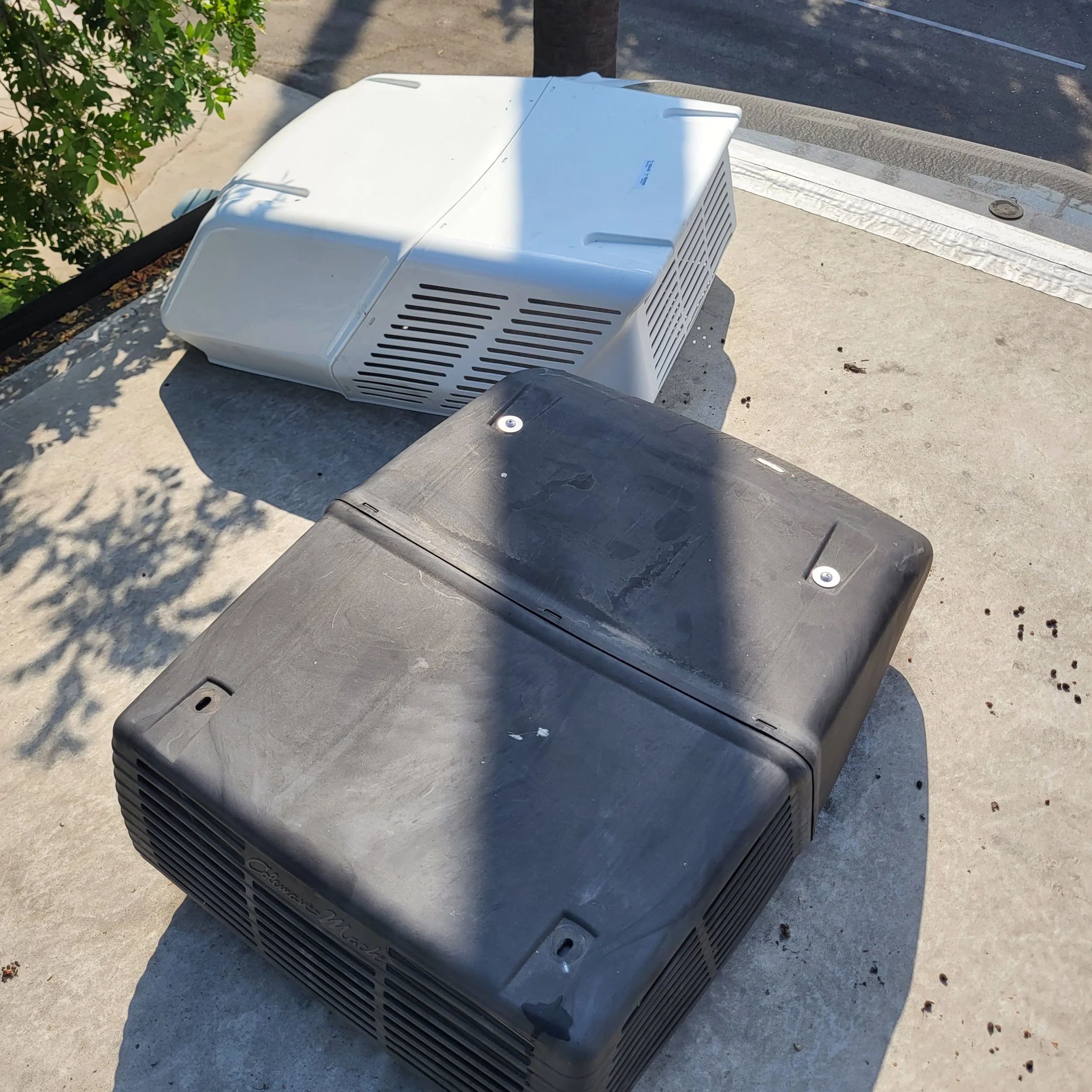 Two air conditioning units, one white and one black, placed on a rooftop ledge with shadows of nearby objects.