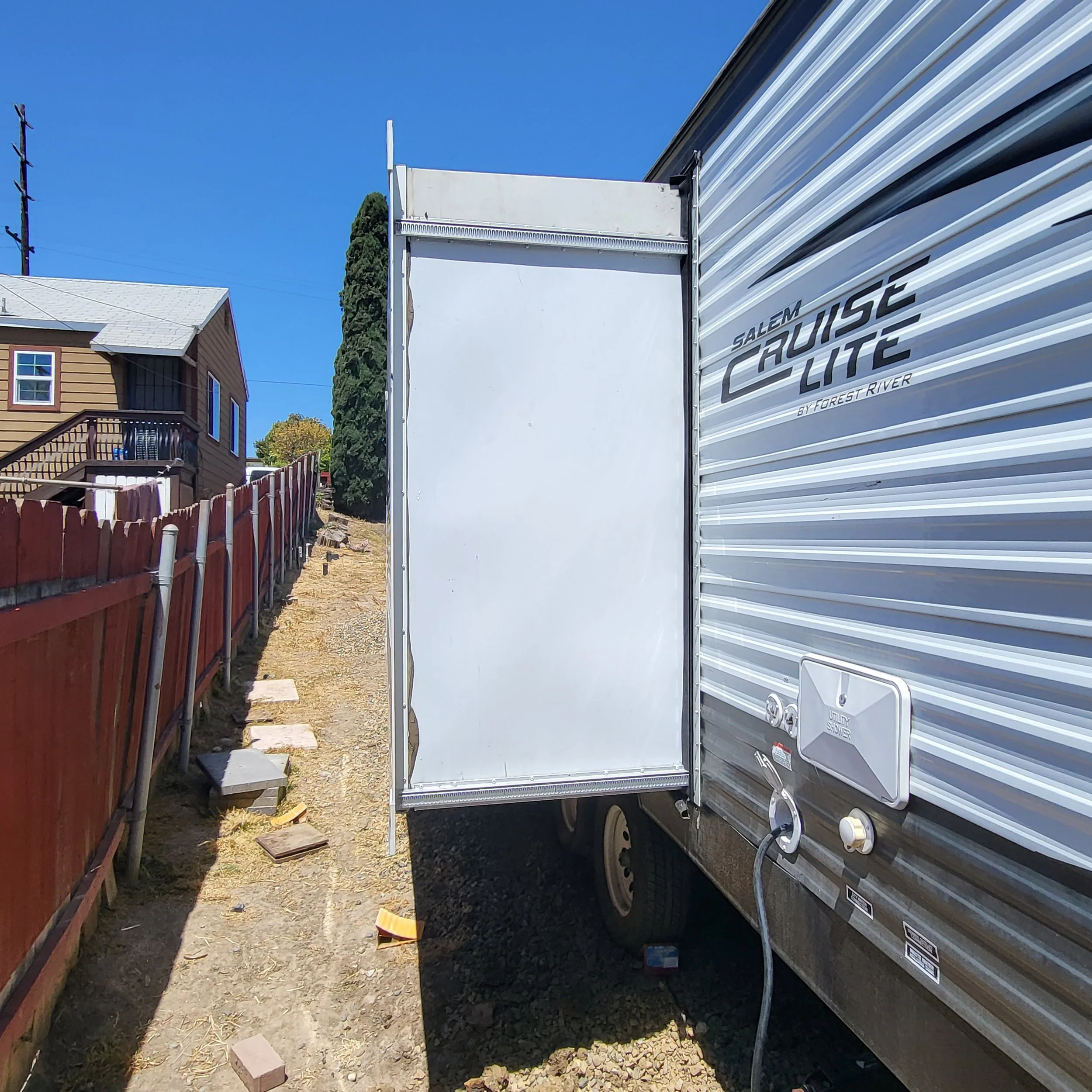 Side view of a Salem Cruise Lite travel trailer, showing the closed back door, electrical hookups, and a dirt pathway with a red wooden fence on the left and a house in the background under a clear blue sky.