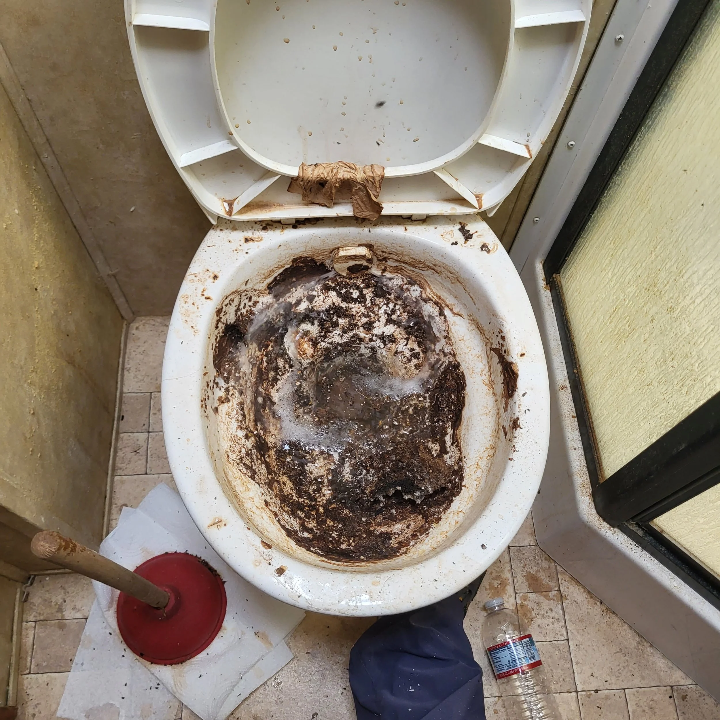 A severely dirty and clogged toilet with brown stains and grime inside the bowl. The seat is lifted, and the surrounding area has cleaning supplies, including a plunger, a spray bottle, and paper towels.