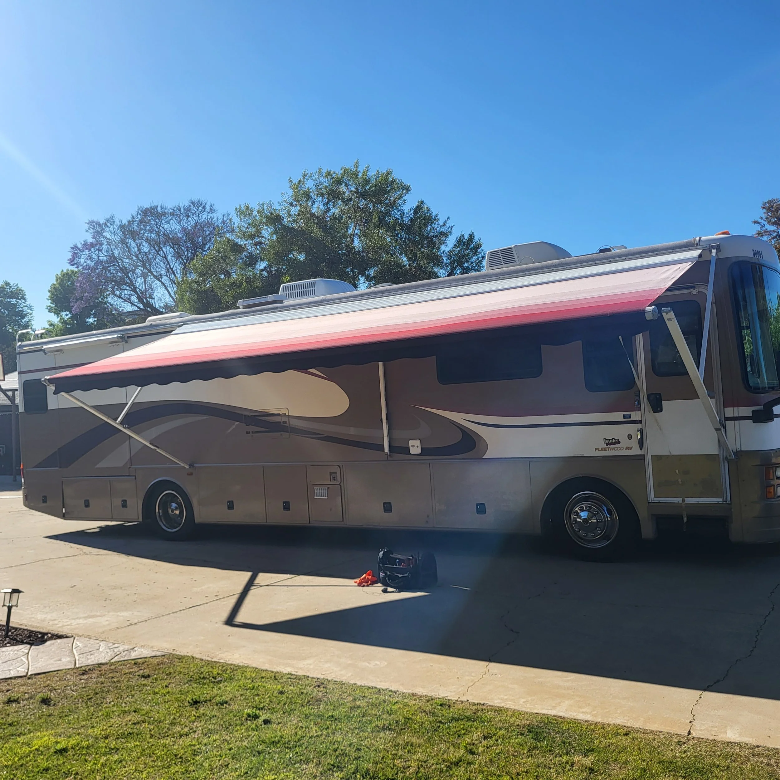 A large motorhome with an extended red and black striped awning, parked on a concrete driveway with grass in the foreground, under a clear blue sky with trees in the background.
