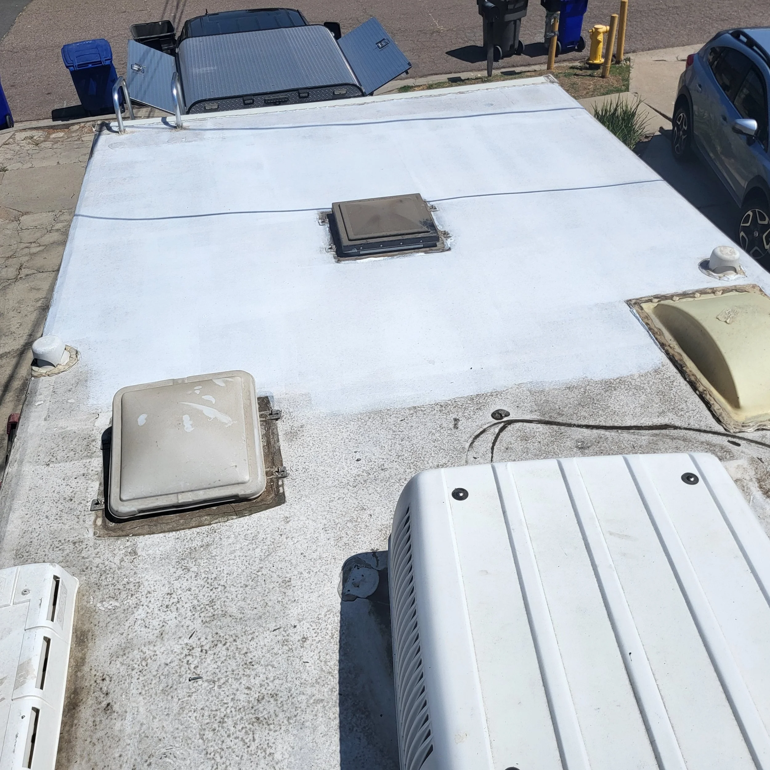 View of the roof of an RV showing vents, an air conditioning unit, and access panels, with nearby cars and recycling bins visible.