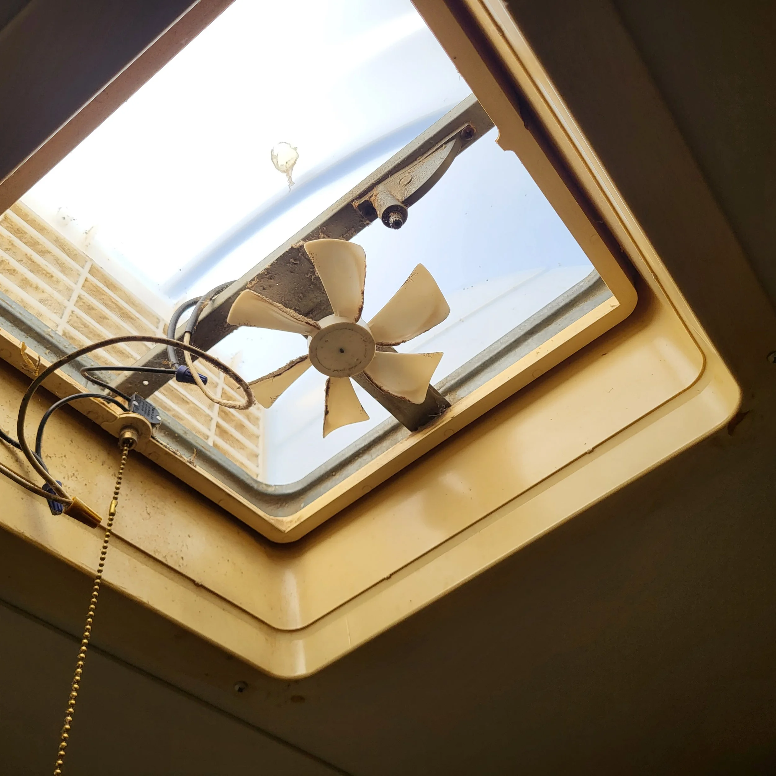 View of an old, broken roof vent with a small fan, in a yellowish frame, seen from inside a building.