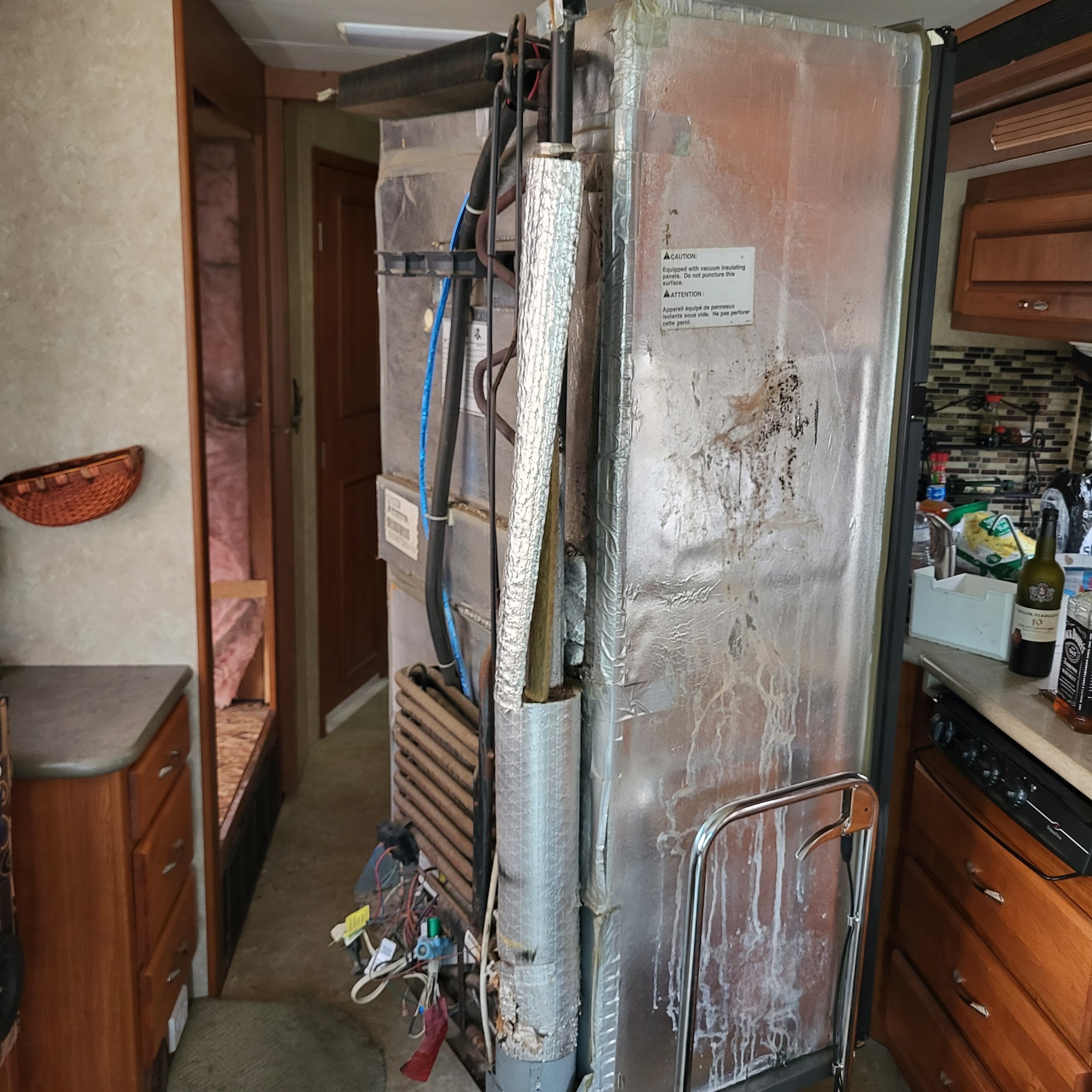 Side view of a refrigerator showing the back panel, wires, and cooling coils in a kitchen with wooden cabinets and a countertop.