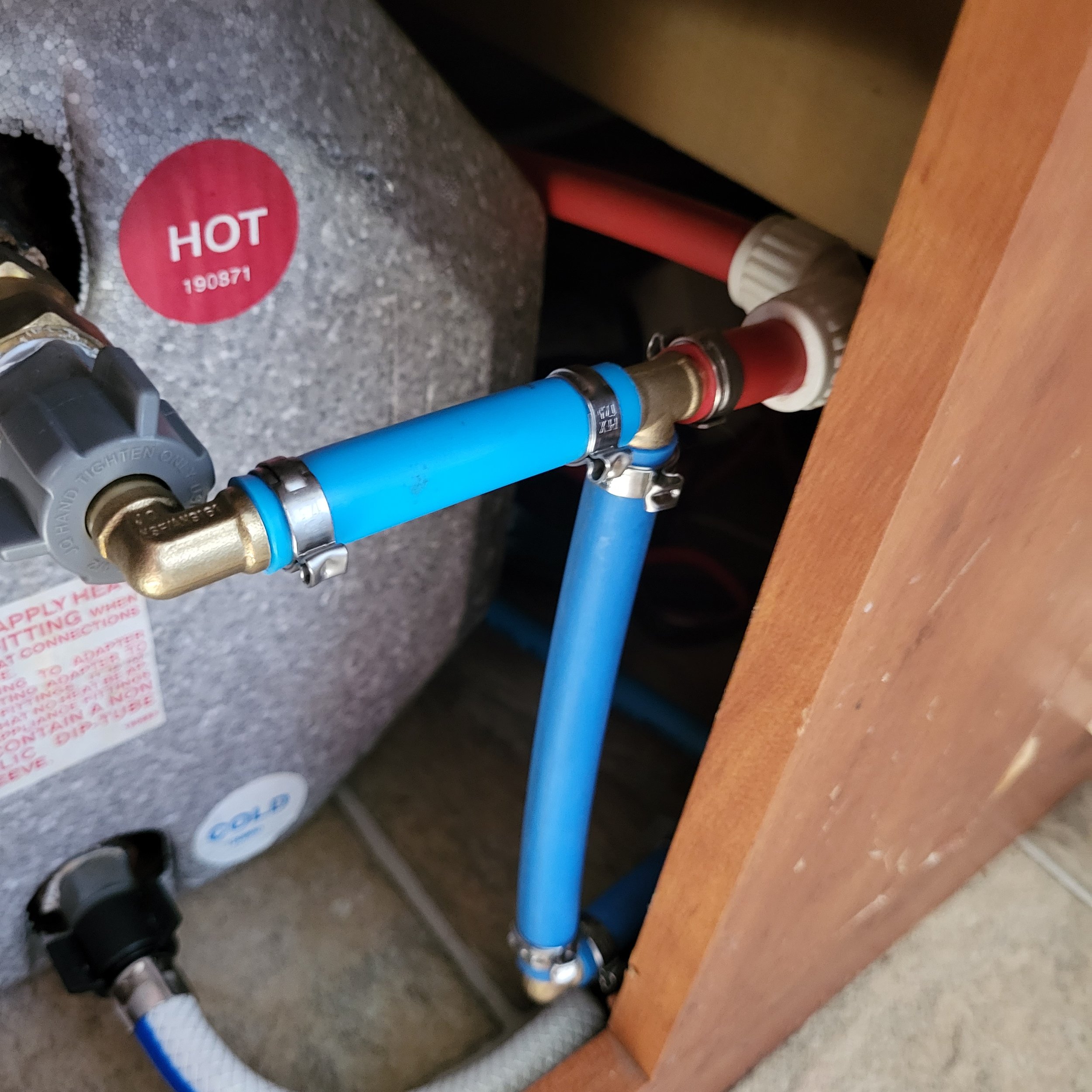 Close-up of plumbing pipes, including a blue pipe, a brass fitting, and a red shut-off valve, connected to a water heater labeled 'HOT' with a red sticker.